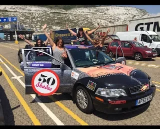 Group of people enjoying a ride in a small, colorful car decorated with various stickers and a '50 Shades' logo, in a parking lot with other cars and a concrete wall in the background.