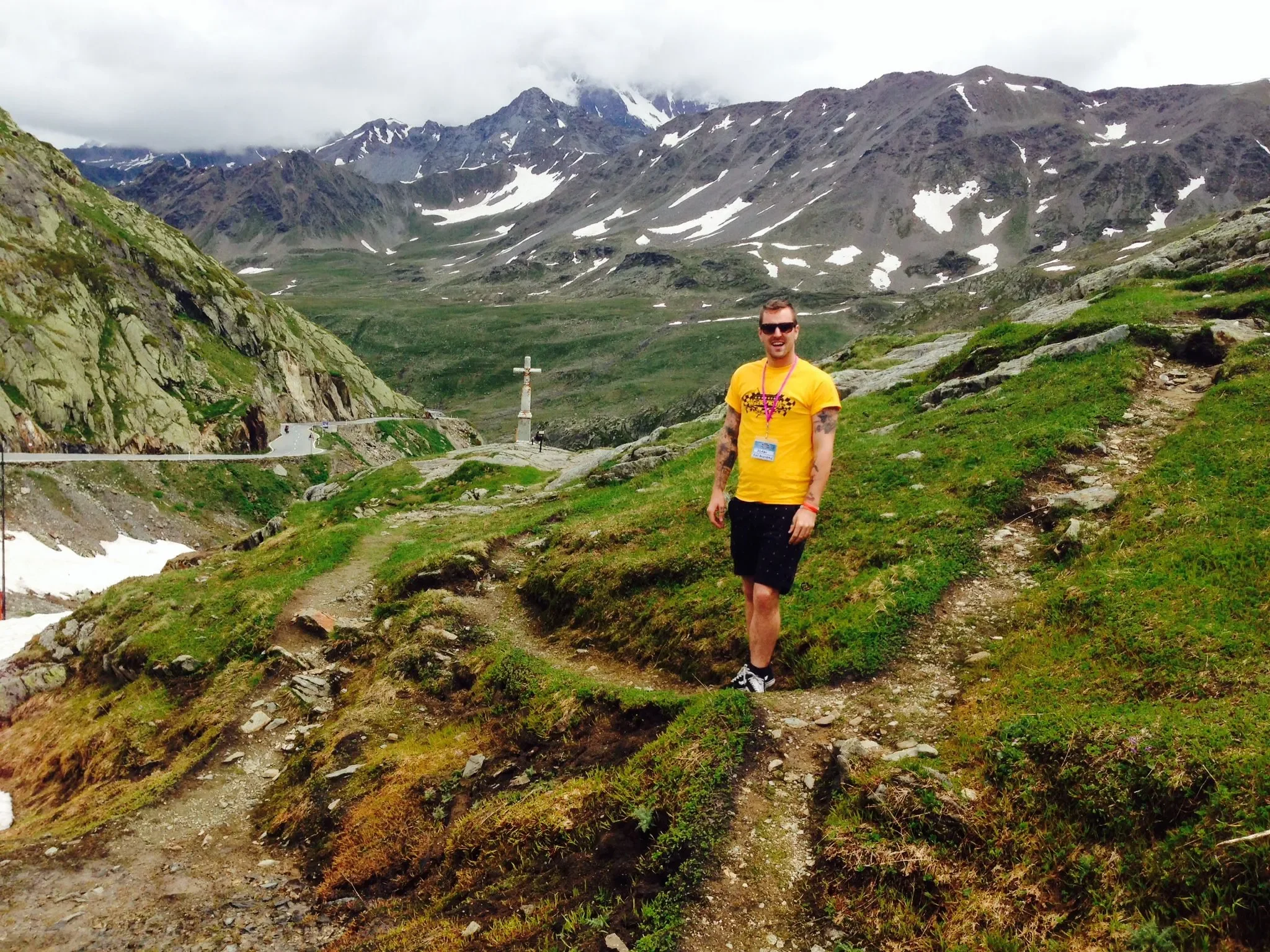 A young man wearing a yellow shirt, black shorts, and sunglasses standing on a mountain trail with snow patches and a cross monument in the background, with rugged mountains and cloudy sky.