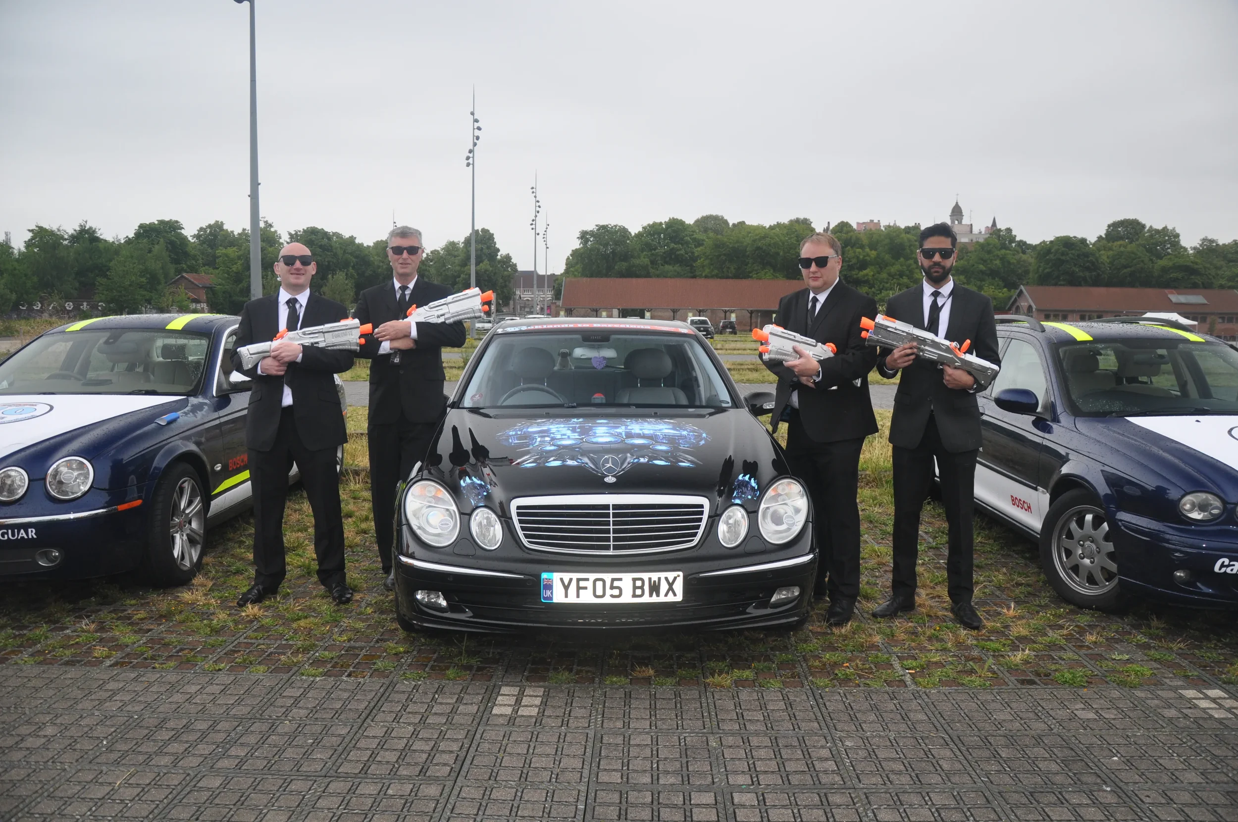 Four men in black suits and sunglasses standing in front of a black Mercedes-Benz car and two police-style cars, holding toy water guns.