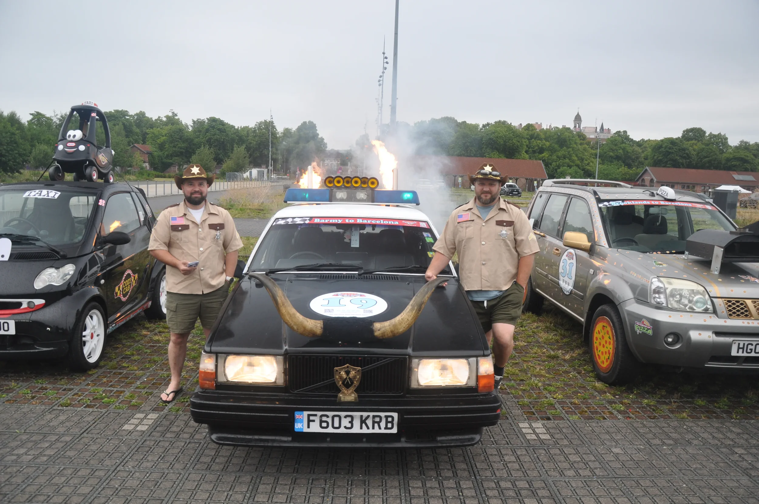 Two men dressed as cowboys standing next to a black taxi with large horns on the hood and a badge on the front, parked between other decorated cars in an outdoor lot with cloudy sky and buildings in the background.