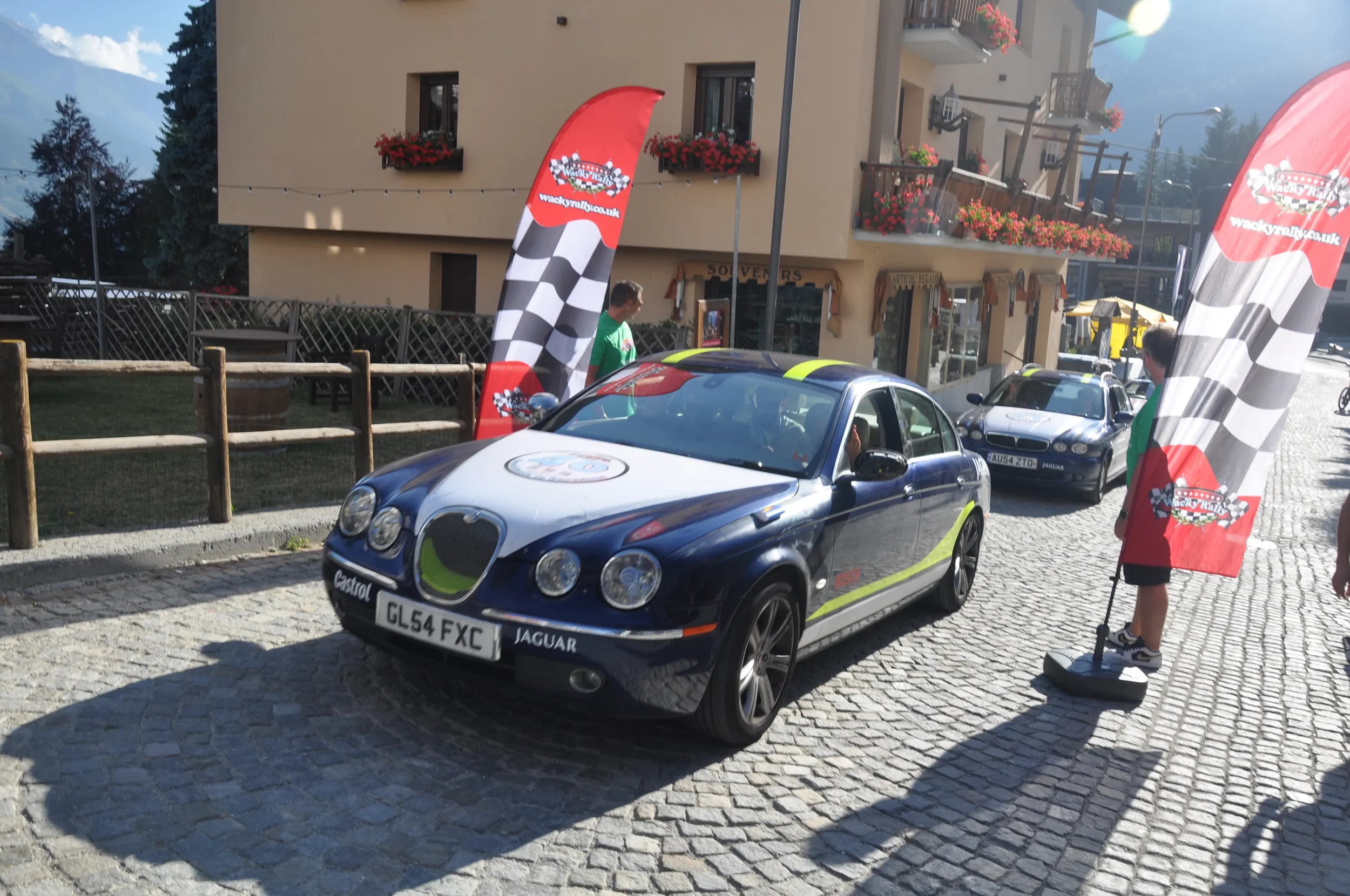 A blue and green Jaguar racing car displayed outdoors, with people and promotional flags around, on a cobblestone street in a mountainous area.