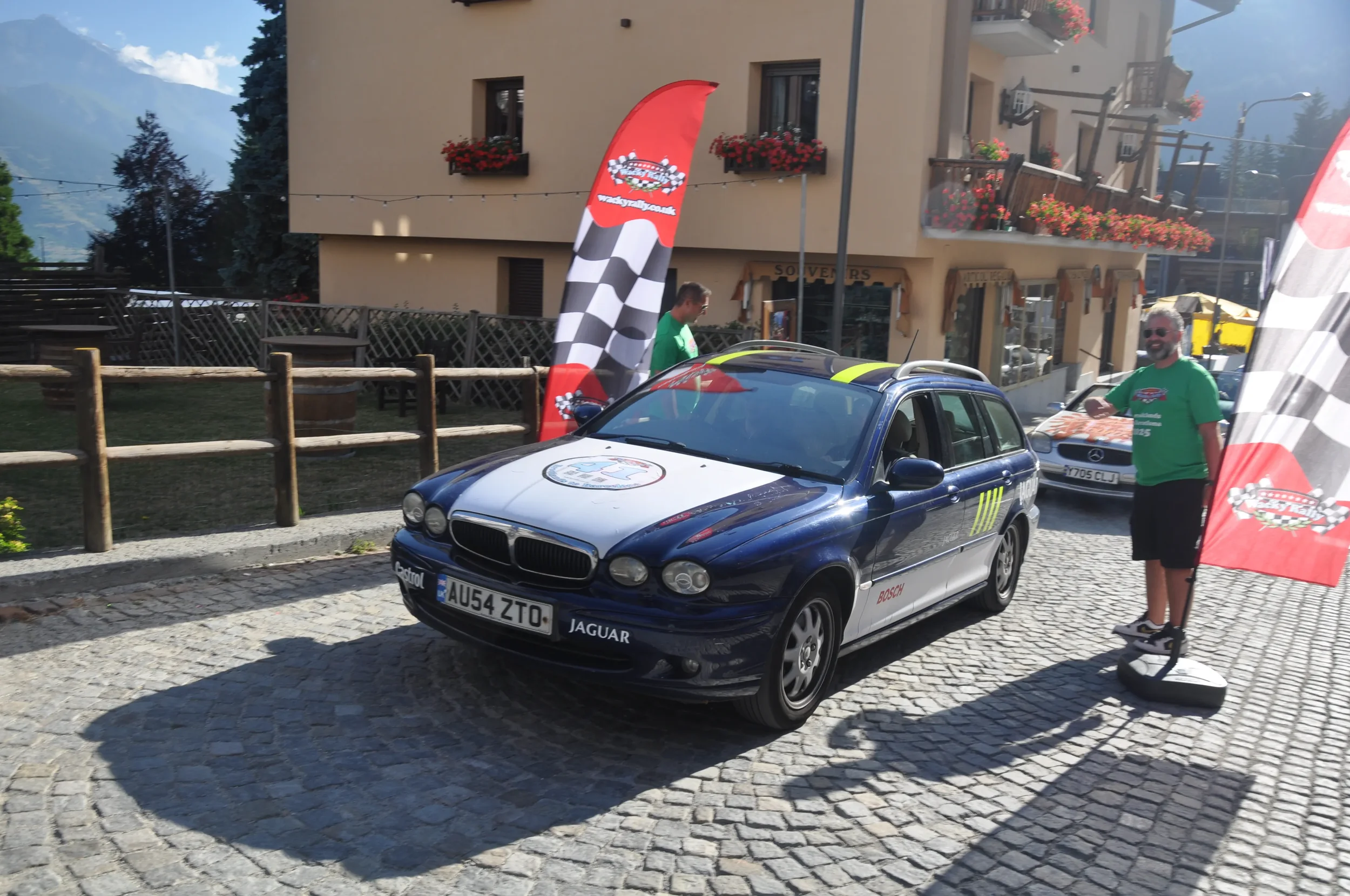 A vintage BMW car with racing decals parked on a cobblestone street during an outdoor event, with people in green shirts and flags with checkered patterns and car logos nearby.