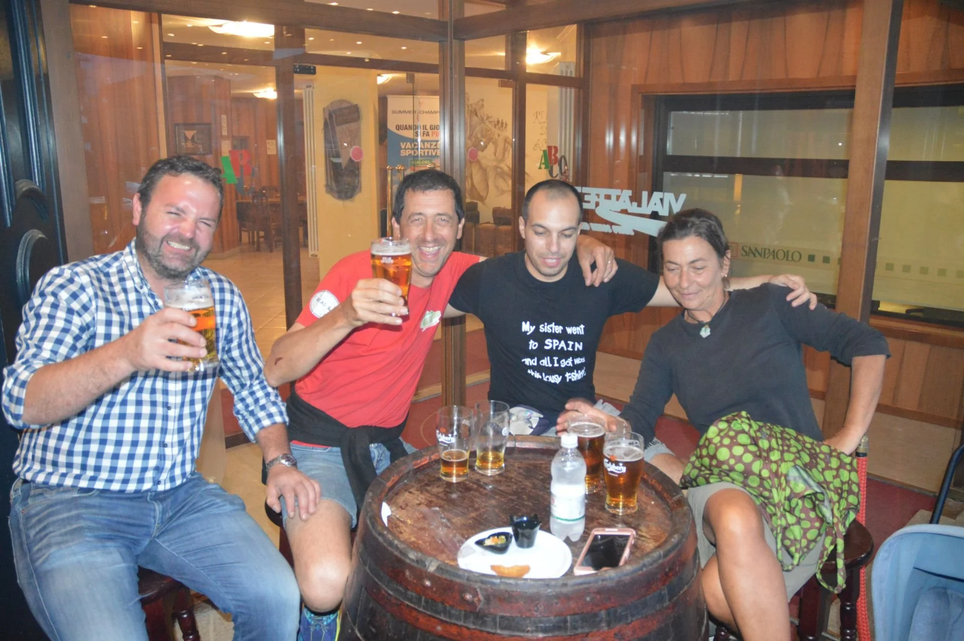 Four friends sitting around a barrel table at a restaurant, holding glasses of beer, smiling, and enjoying a social gathering.