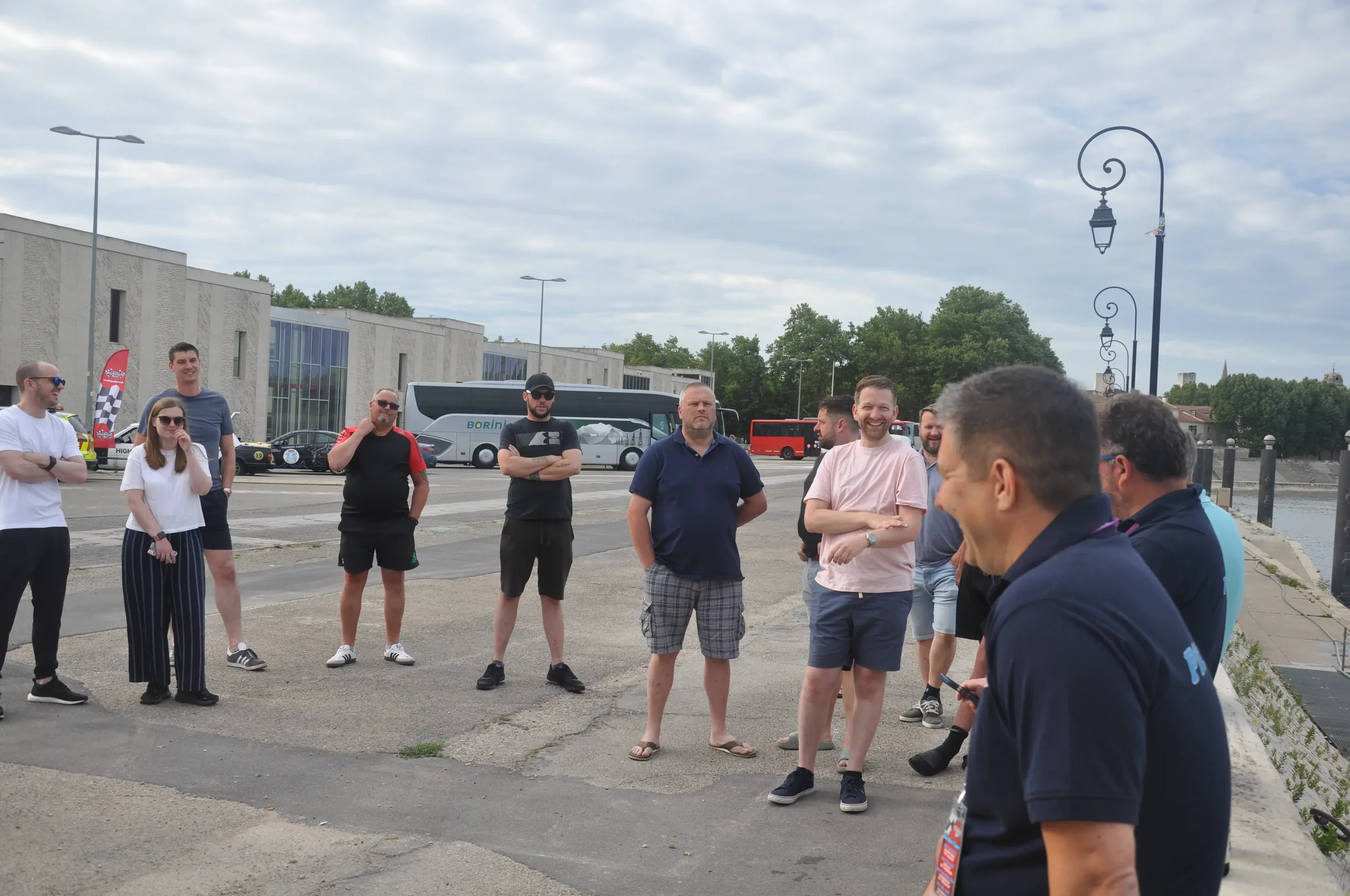 A group of people standing outdoors near a body of water, some smiling and talking, with buses and buildings in the background on a cloudy day.
