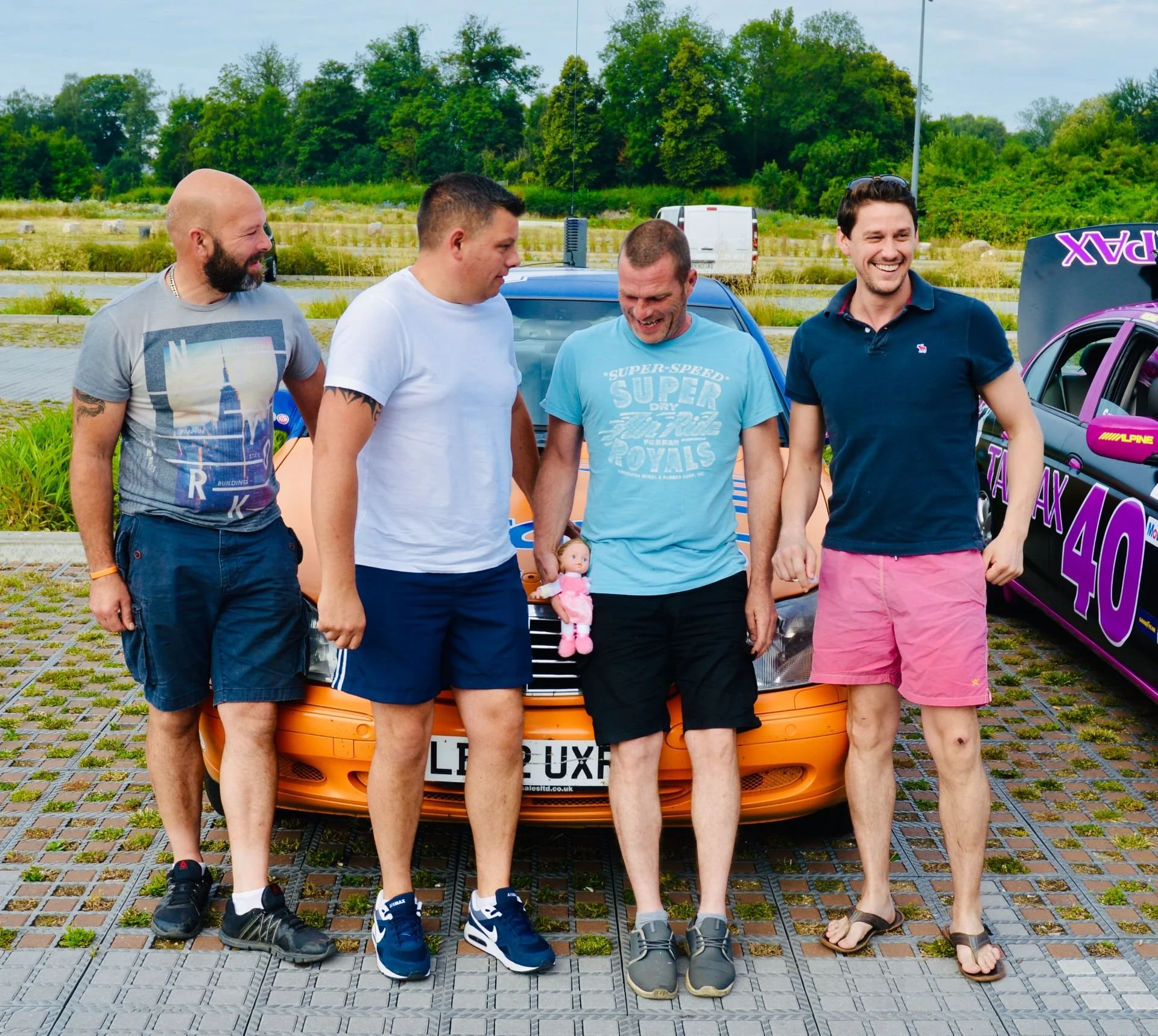 Four men standing in front of a car, smiling and appearing to share a moment of laughter. One man is holding a small doll. The background shows a grassy field and trees, with another sports car to the right.