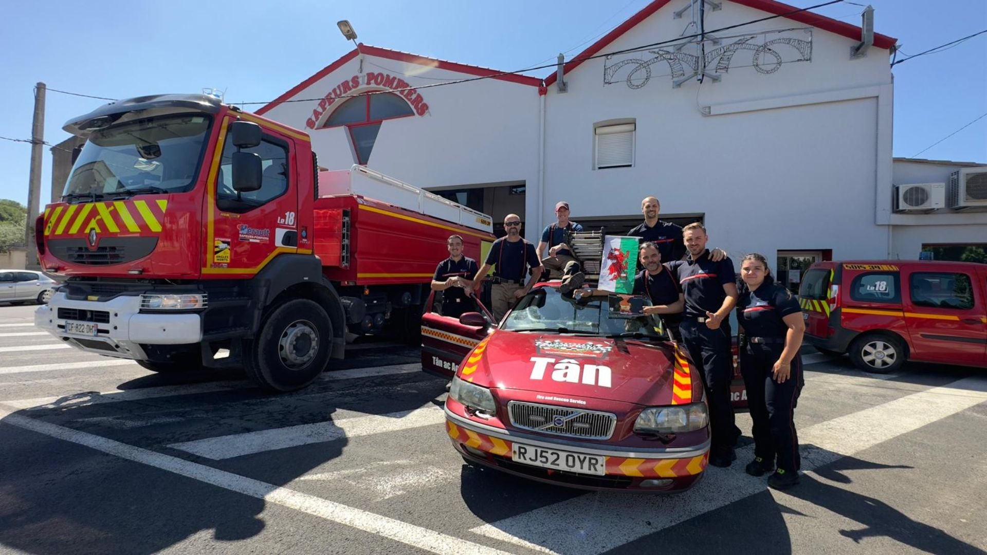 Group of firefighters and emergency responders with a fire truck and a patrol car in front of fire station building, holding equipment and a small flag.