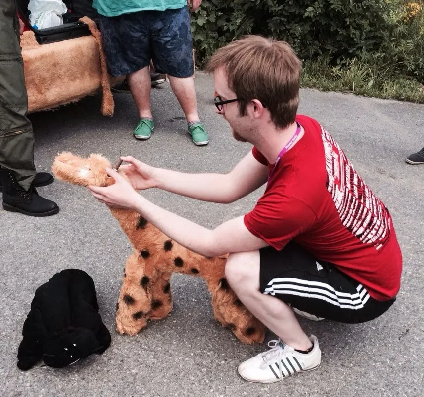 A young man in a red t-shirt and black shorts is kneeling on the ground, fitting a plush giraffe costume onto a person or mannequin. The setting appears to be outdoors, with other people and greenery in the background.