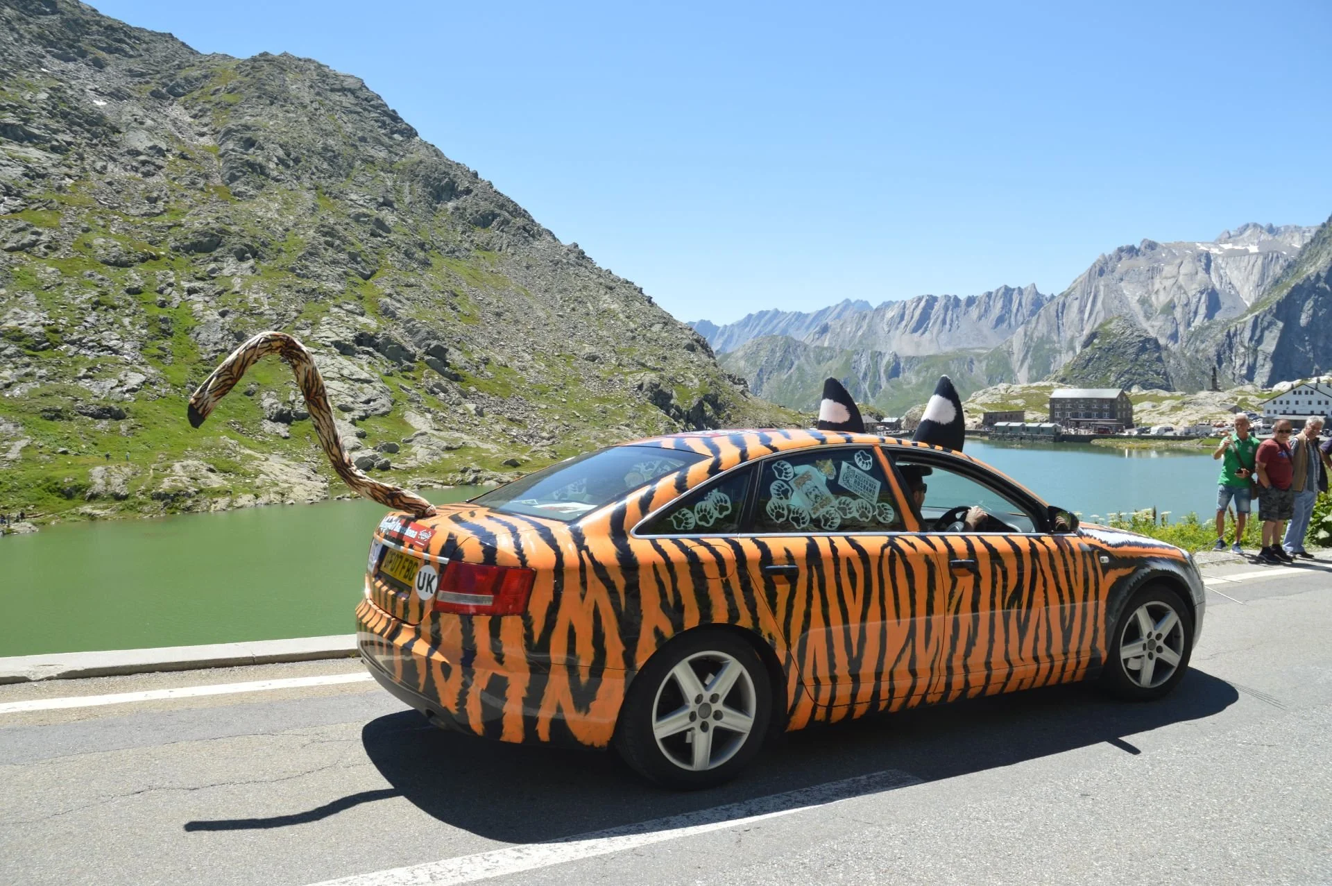 A car decorated with a tiger stripe pattern, featuring tiger ears, a tail, and tiger face details on the front, parked by a mountain lake with rugged mountains and a small building in the background, and three people standing nearby.