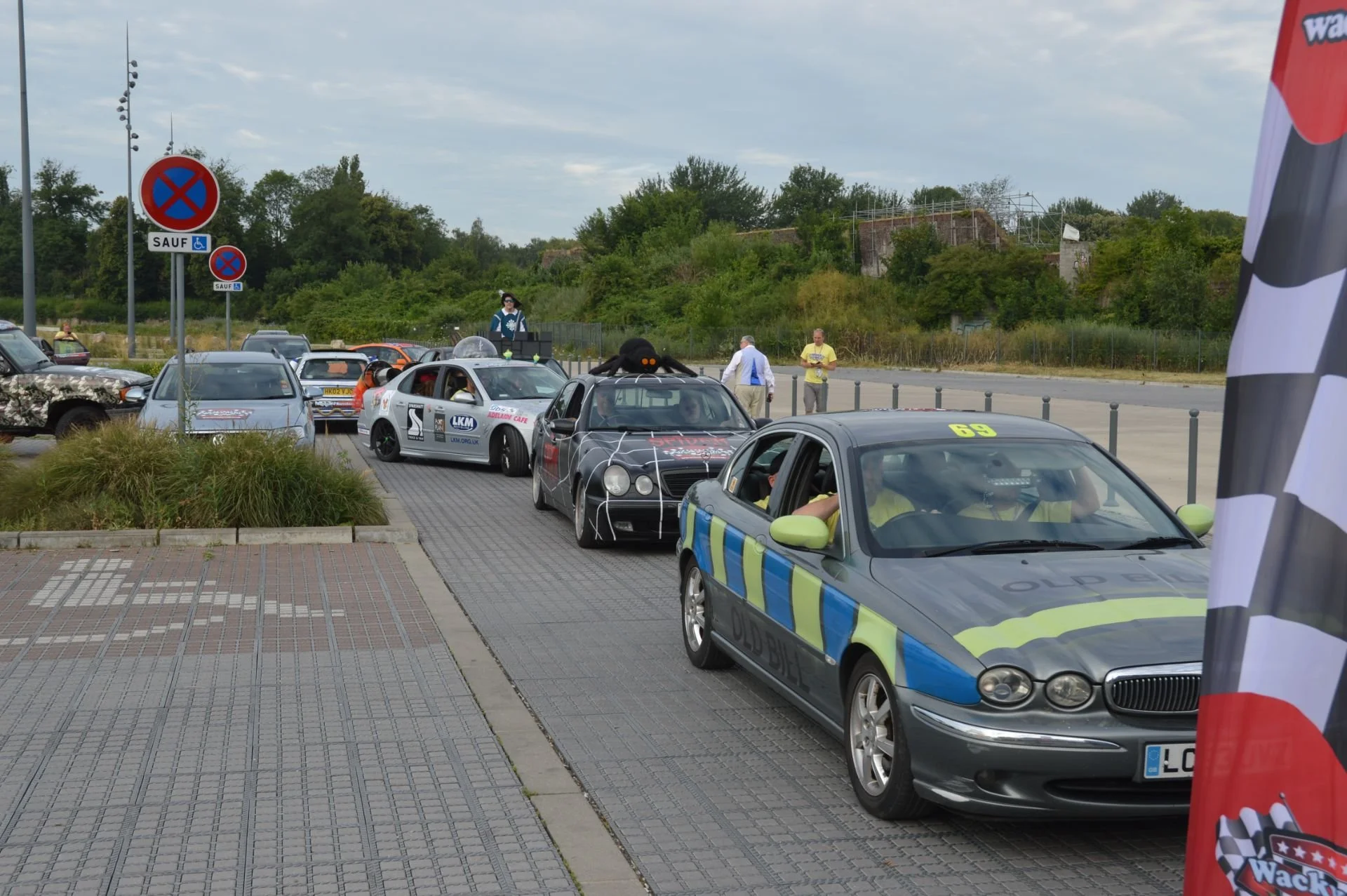 Line of decorated cars, including an old police car with a checkered pattern, parked on a pavement at an outdoor event with a flag on the right side and people standing and observing in the background.