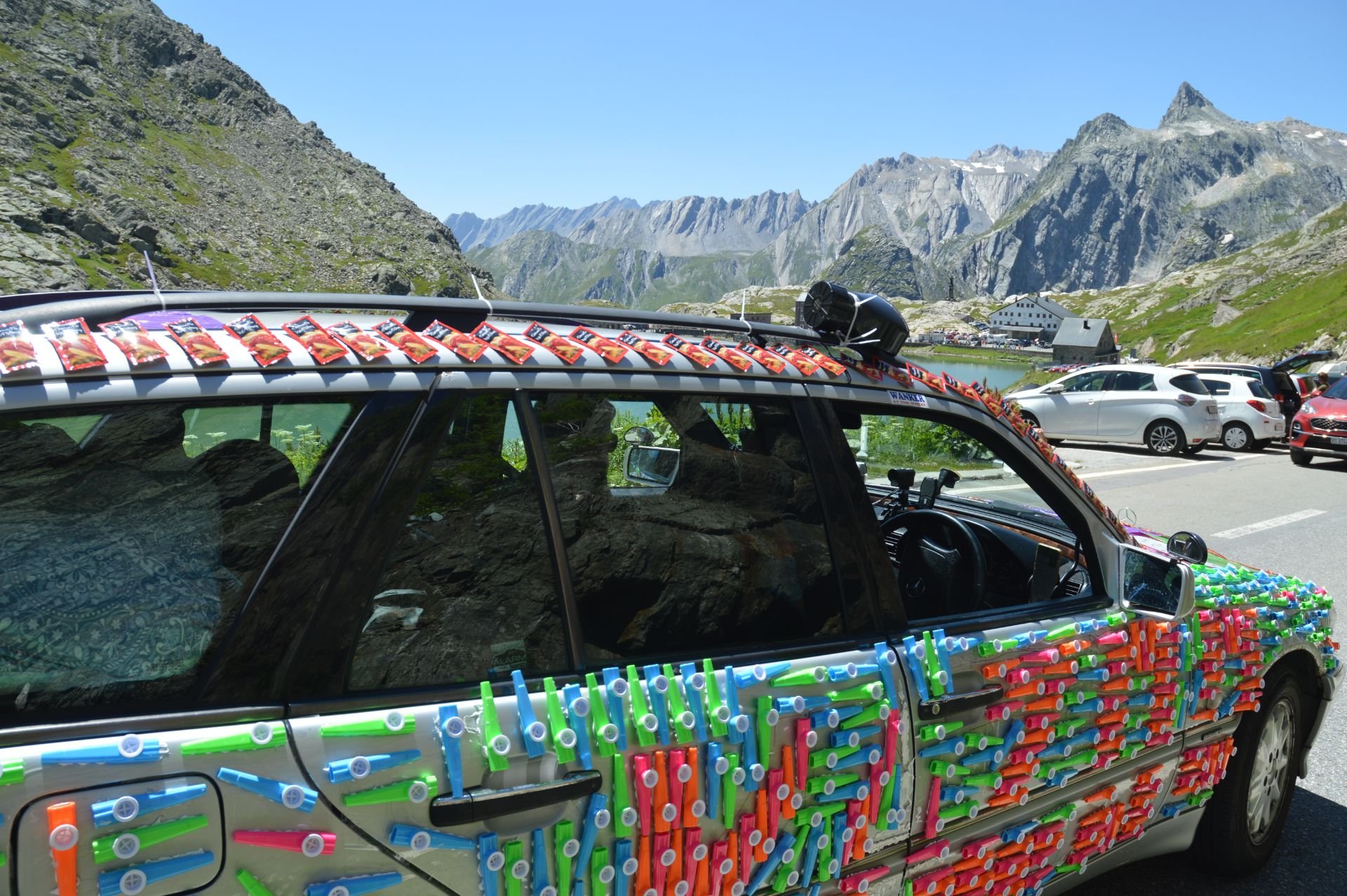 Car decorated with numerous colorful plastic straws in a mountainous area with parked cars and lakes.