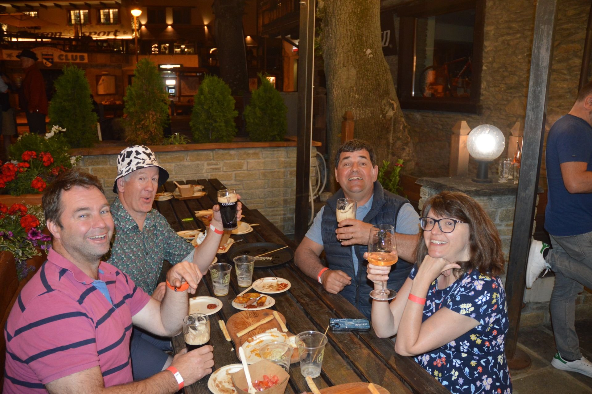 Four people sitting at a wooden table outdoors at night, smiling and holding drinks, with pizza and snacks on the table, in a cozy restaurant setting with plants, flowers, and decorative lighting.