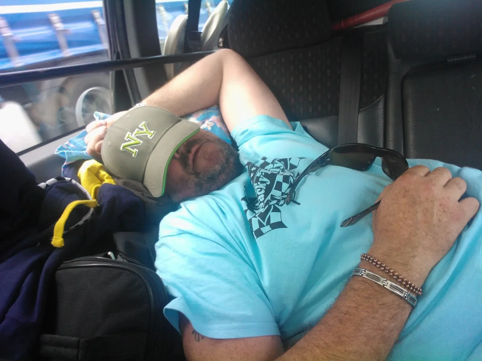 A man with a beard wearing a blue T-shirt, a tan cap, and sunglasses resting on his chest, sleeping on a bus. He has bracelets on his wrist and a tattoo on his arm. There are bags and other items around him, and he is leaning against the window.