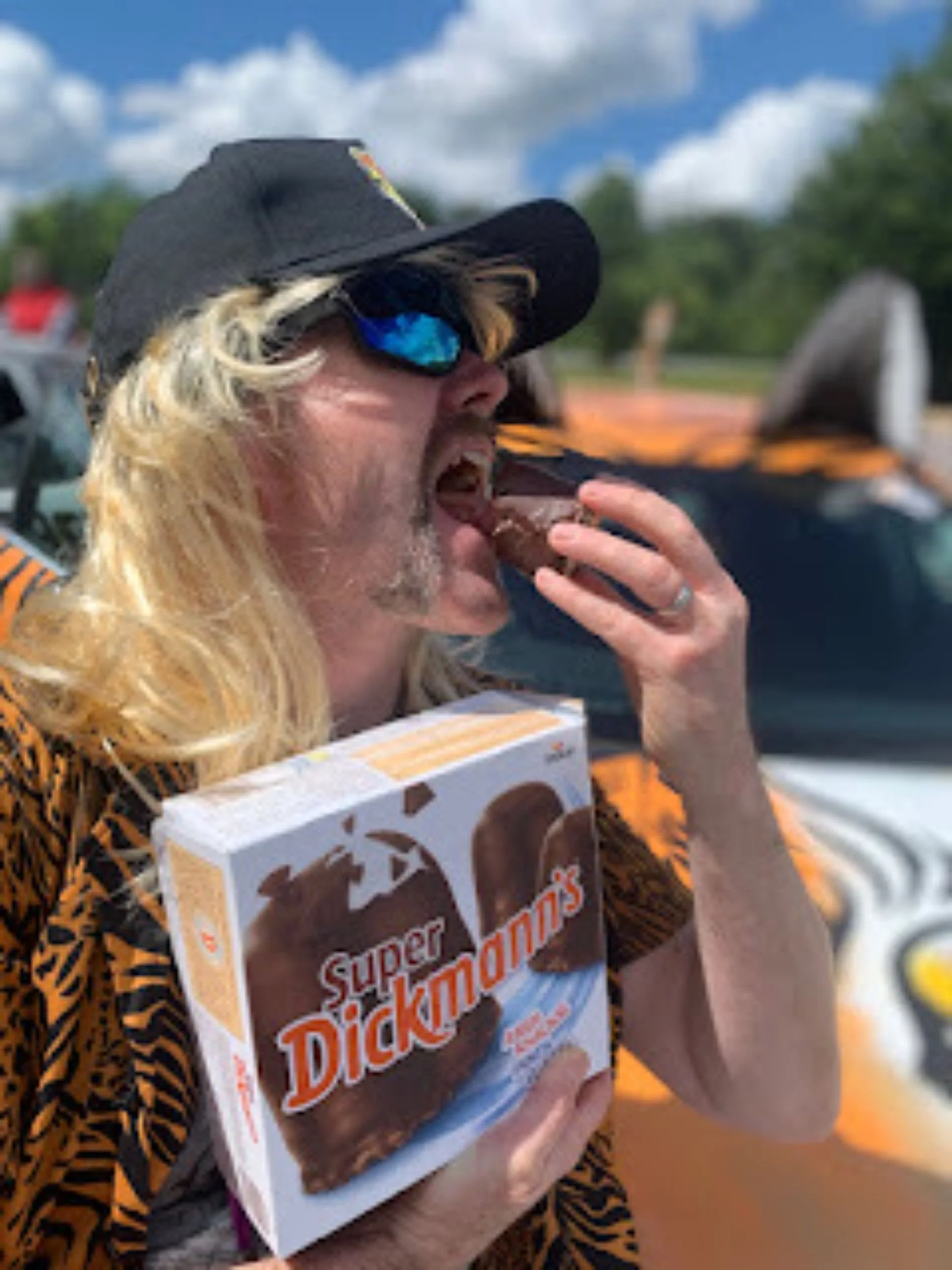 A man with long blond hair, sunglasses, and a black cap eating a chocolate ice cream cone while holding a box of Super Dickmann's chocolates outside on a sunny day.