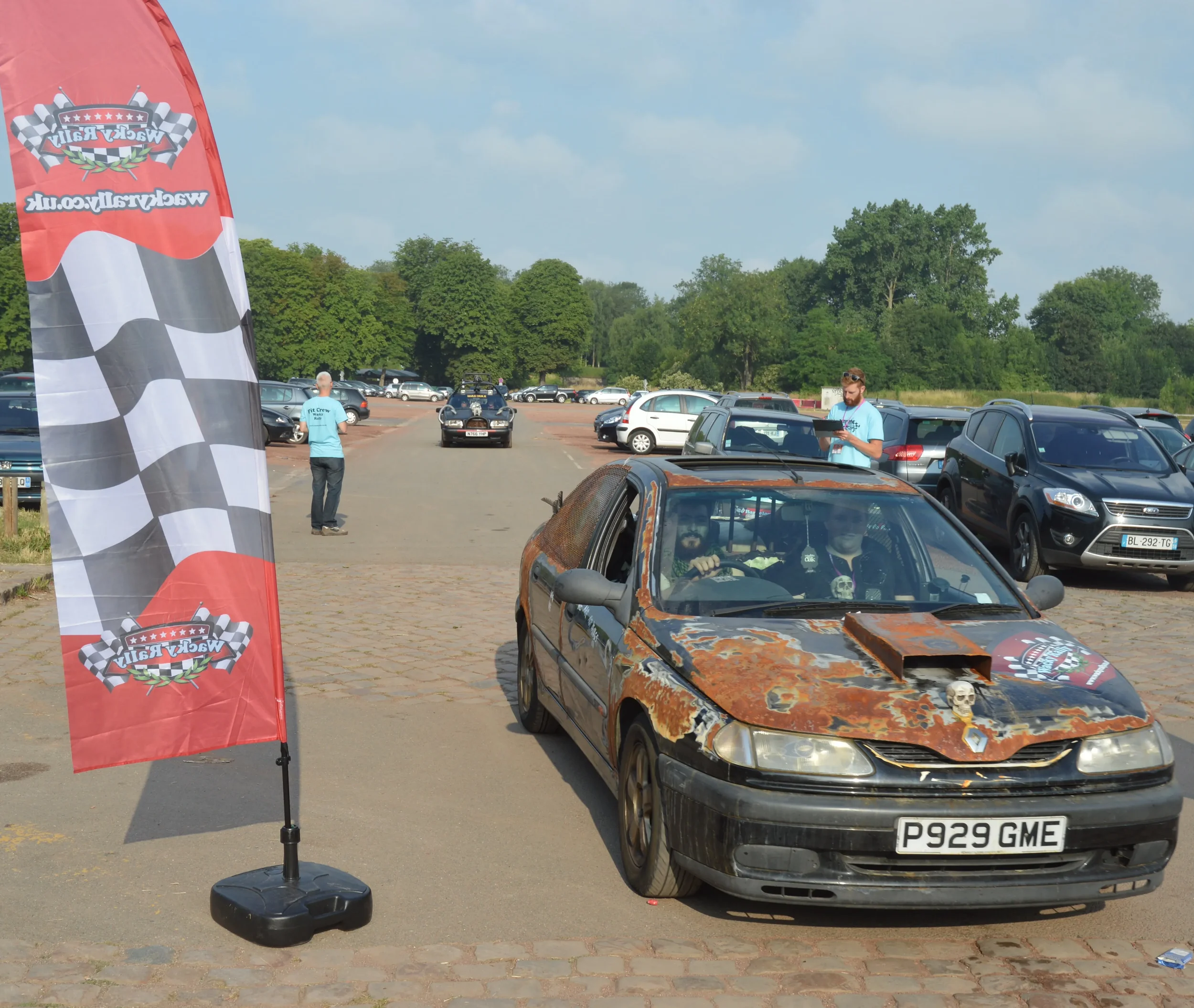 Rusty, weathered car with a skull on its hood, parked in a lot during a racing event, with people around and a checkered flag banner to the side.