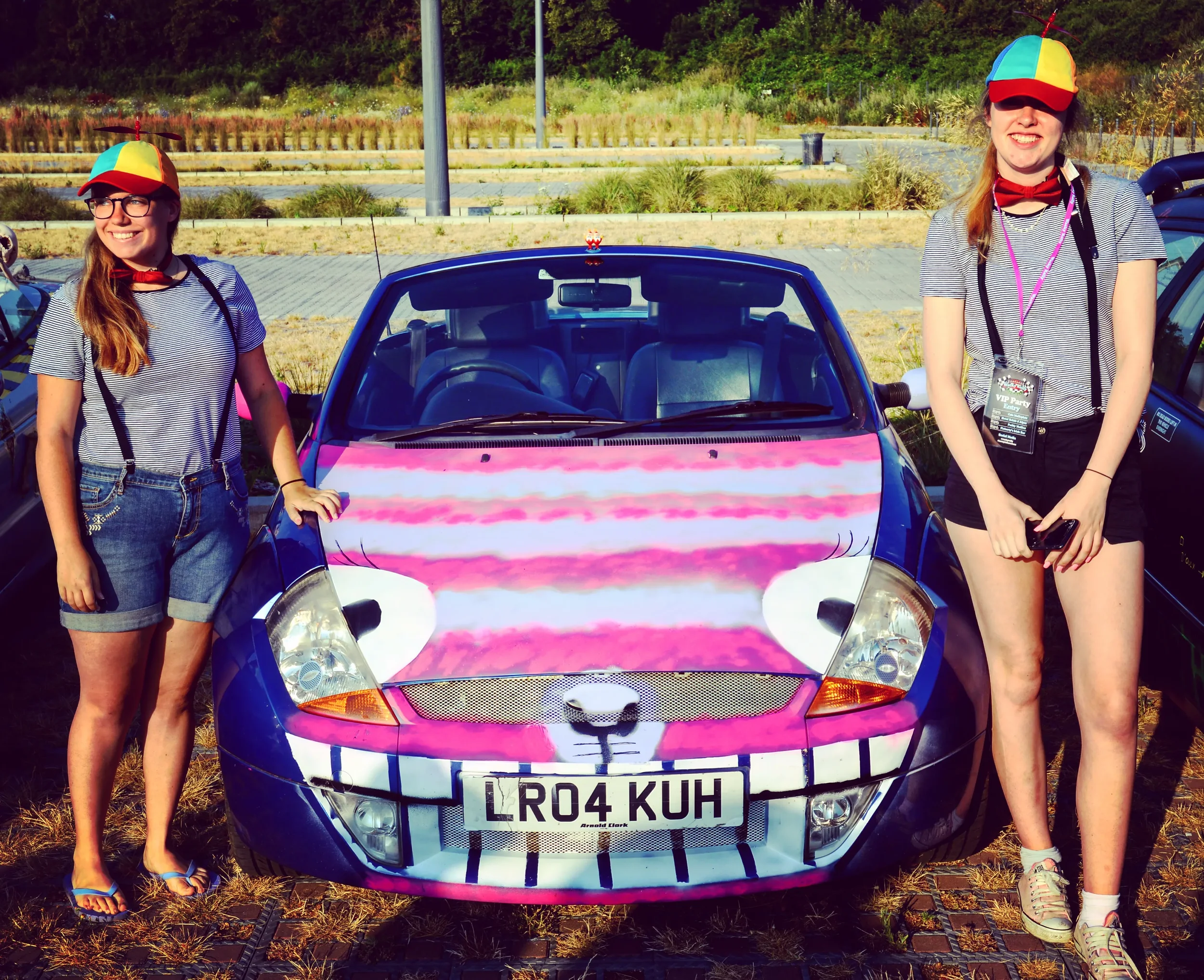 Two young women standing on either side of a small car with pink and white race stripes, wearing striped t-shirts, shorts, and colorful hats, smiling outdoors on a sunny day.