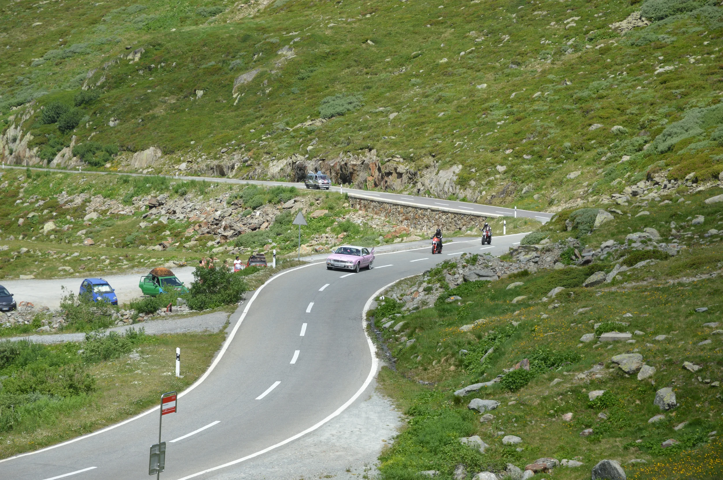Scenic mountain road with cars and motorcycles traveling through green, rocky landscape.