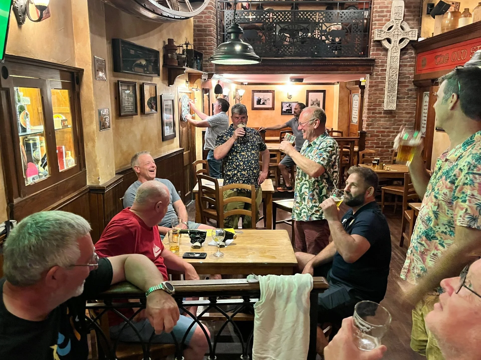 Group of men socializing and enjoying drinks in a cozy pub, with some standing and others sitting at tables, decorated with framed pictures and a large Celtic cross.