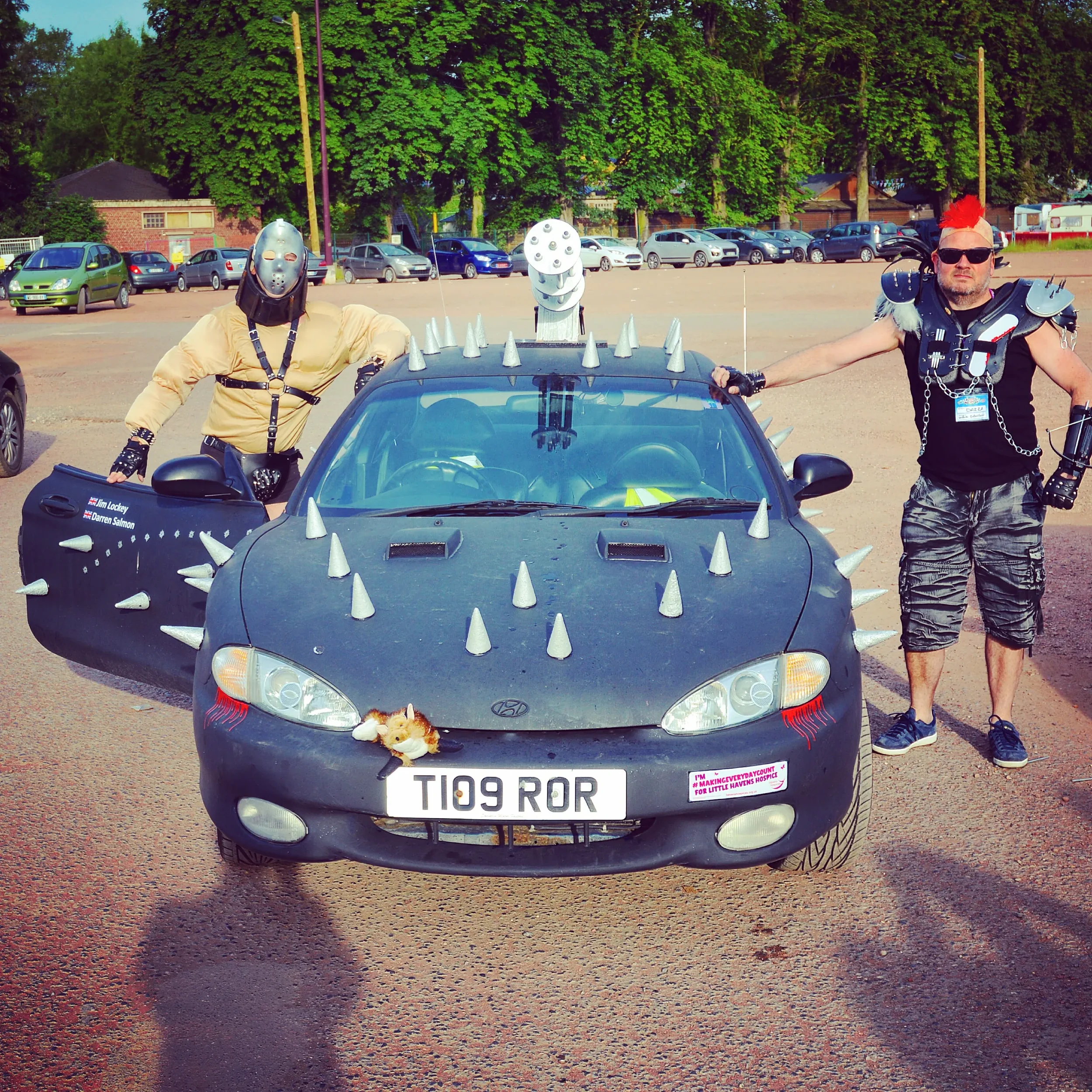Two men dressed in punk and sci-fi costumes posing beside a customized black car decorated with spikes and a stuffed animal on the front bumper. The car has a unique license plate reading 'T109 ROR' and appears to be at a public event or gathering.