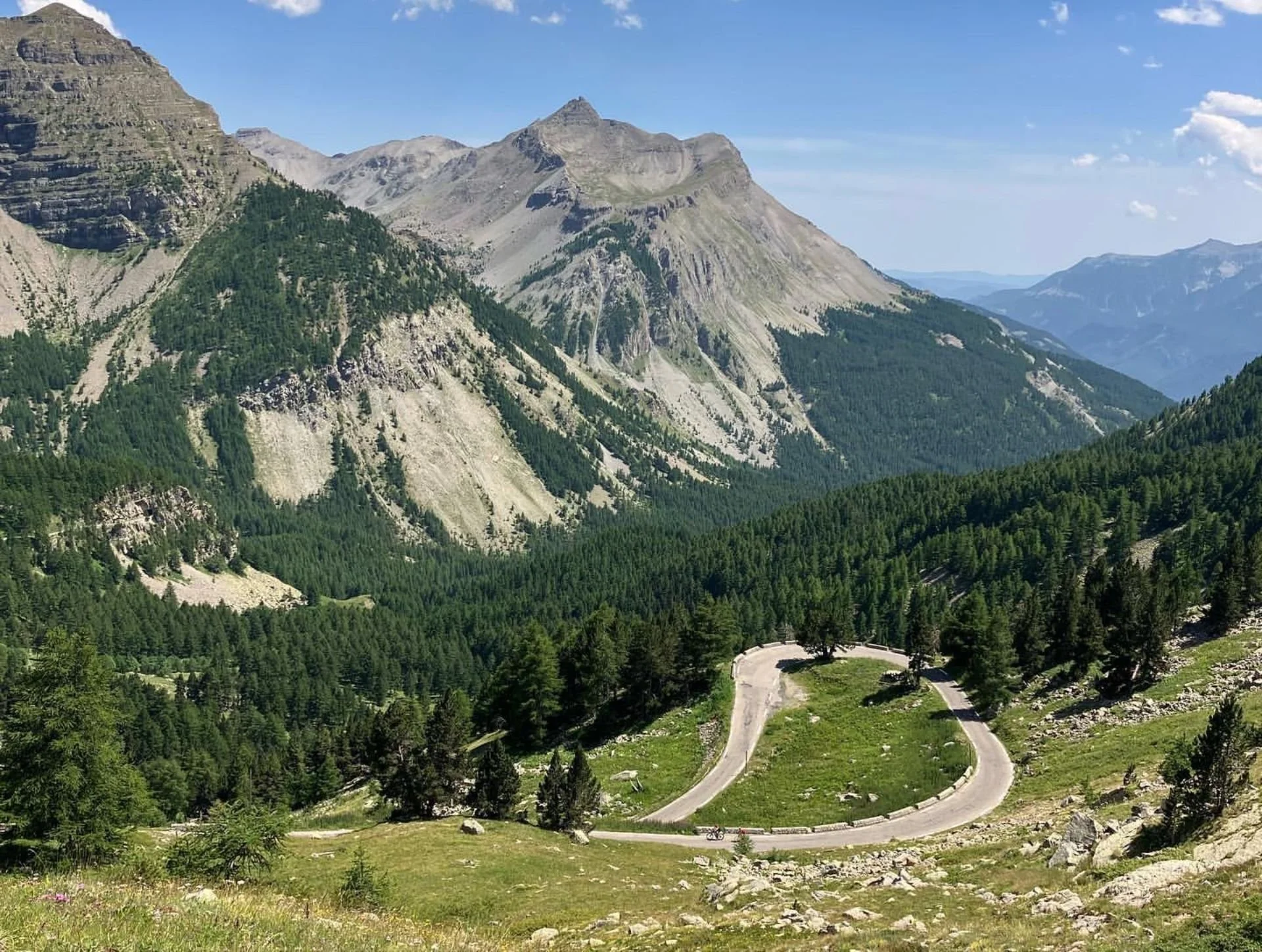 Mountain landscape with winding road, dense green forest, and tall rocky peaks under a blue sky with clouds.