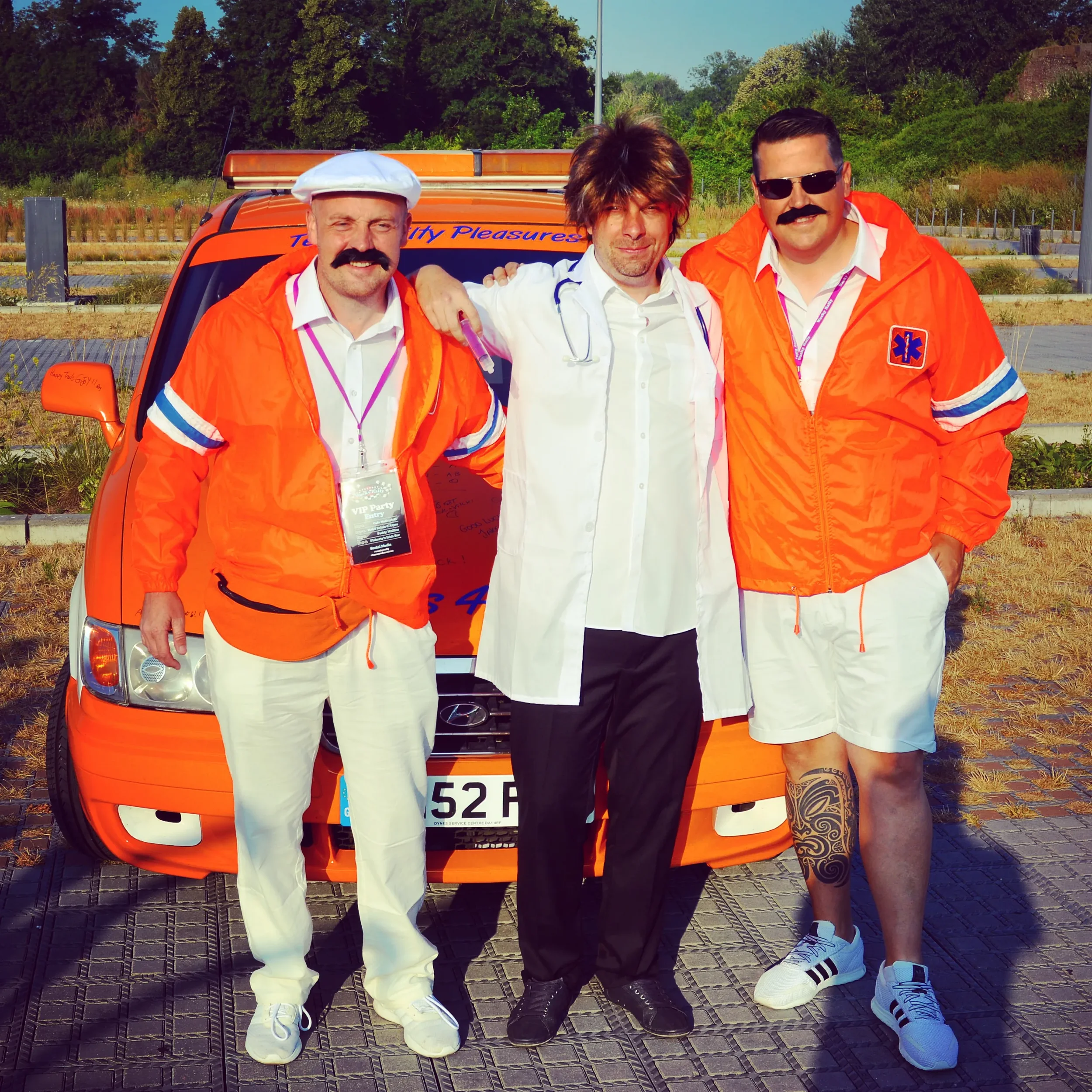 Three men dressed as medical professionals or emergency workers standing in front of an orange vehicle. The two men on the left and right wear orange jackets with medical symbols and white shorts, while the man in the middle wears a white medical coa
