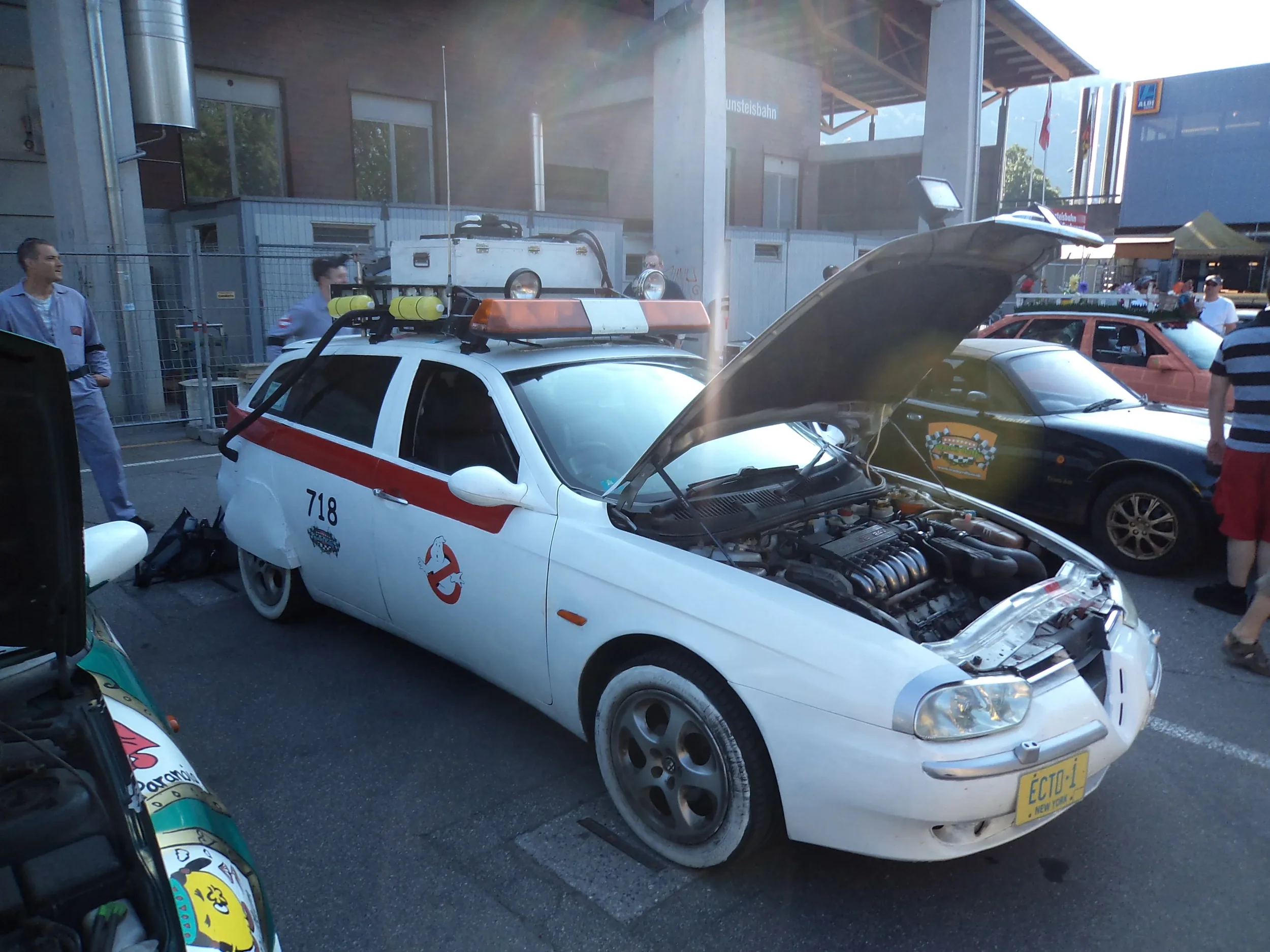 A white replica of the Ecto-1 from Ghostbusters with the hood open, parked at a car show. The vehicle has Ghostbusters logos and is surrounded by people and other cars.