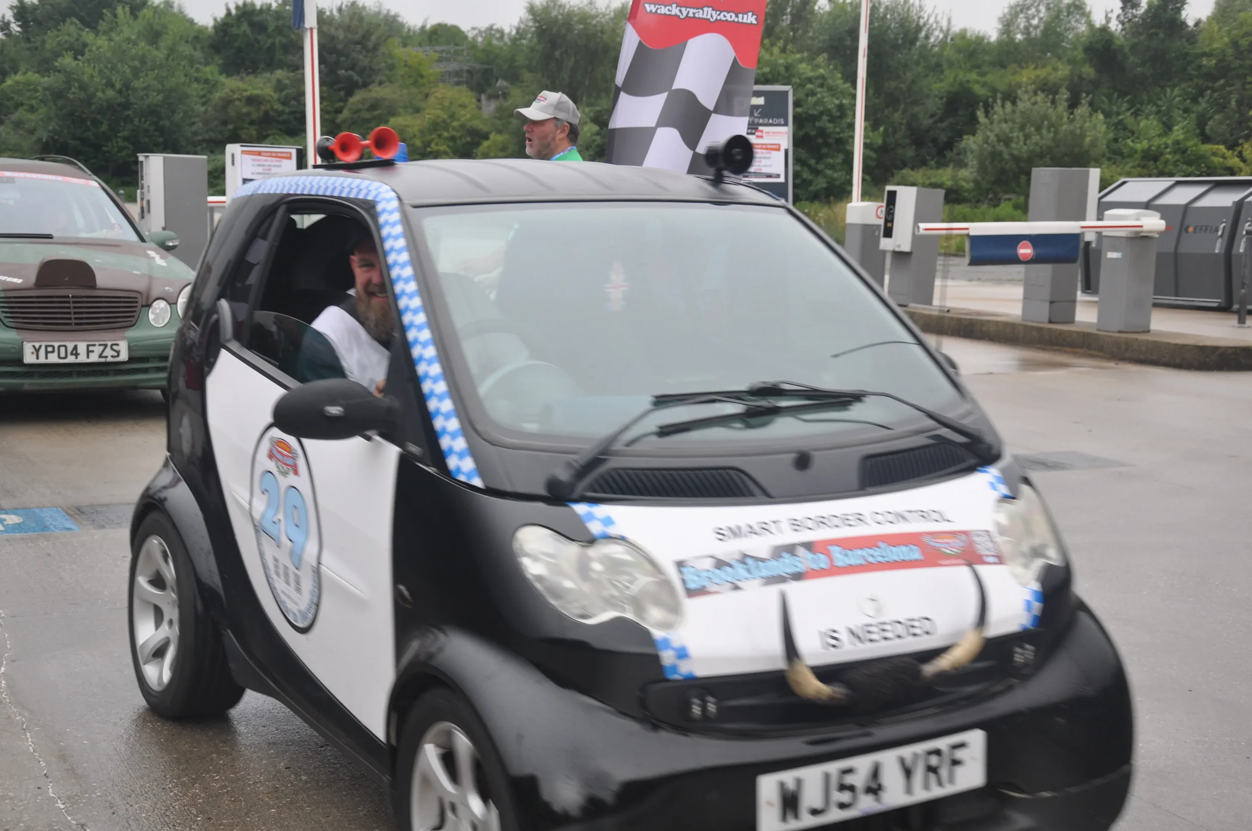 A small black and white electric car with the number 29 on the side, decorated with blue checkered trim, and a sign reading "IS NEEDED" on the front. The car is participating in a rally race, as indicated by the "Smart Border Control" banner and vari