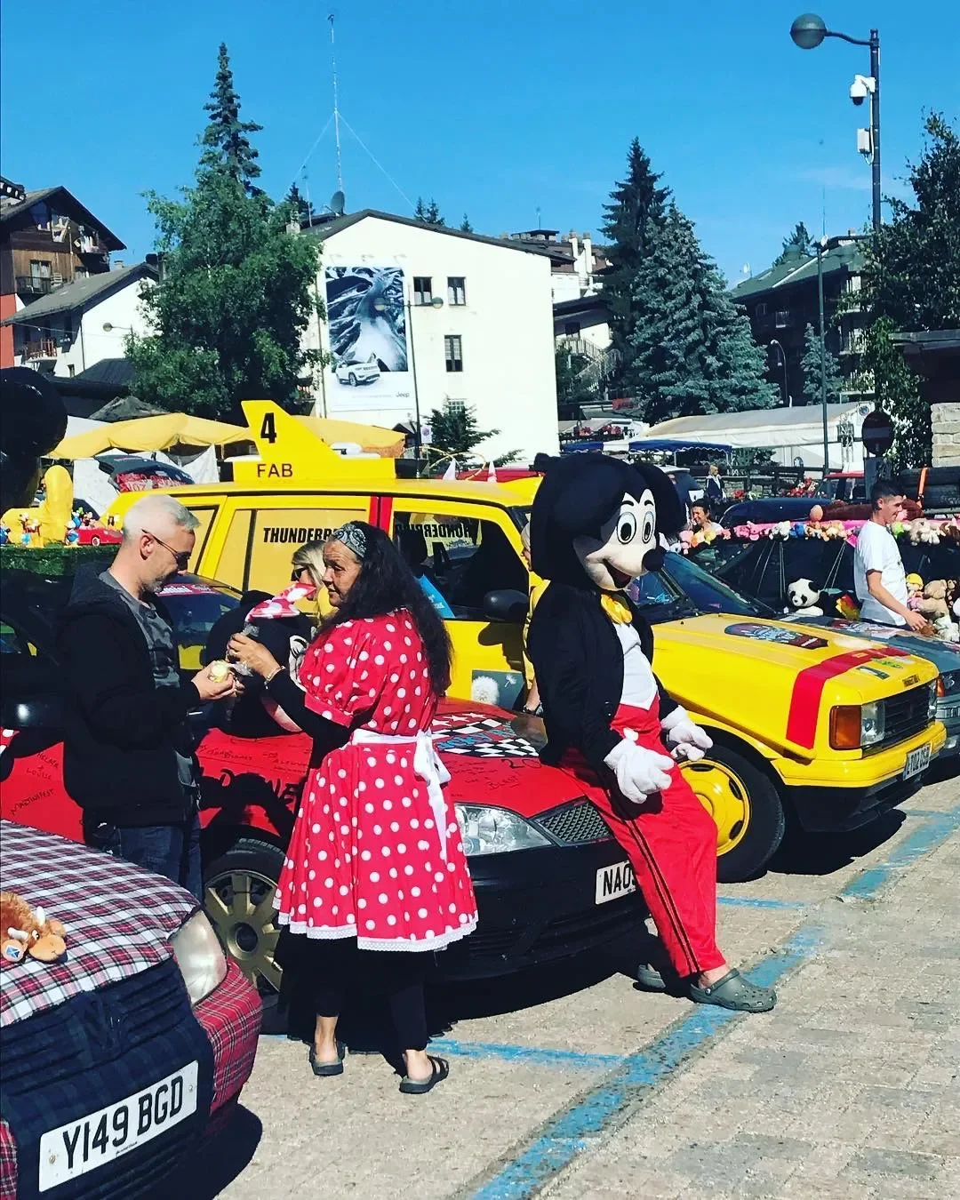 People at a car event with two decorated cars, including a person in a Mickey Mouse costume, and others dressed in costumes or costumes.