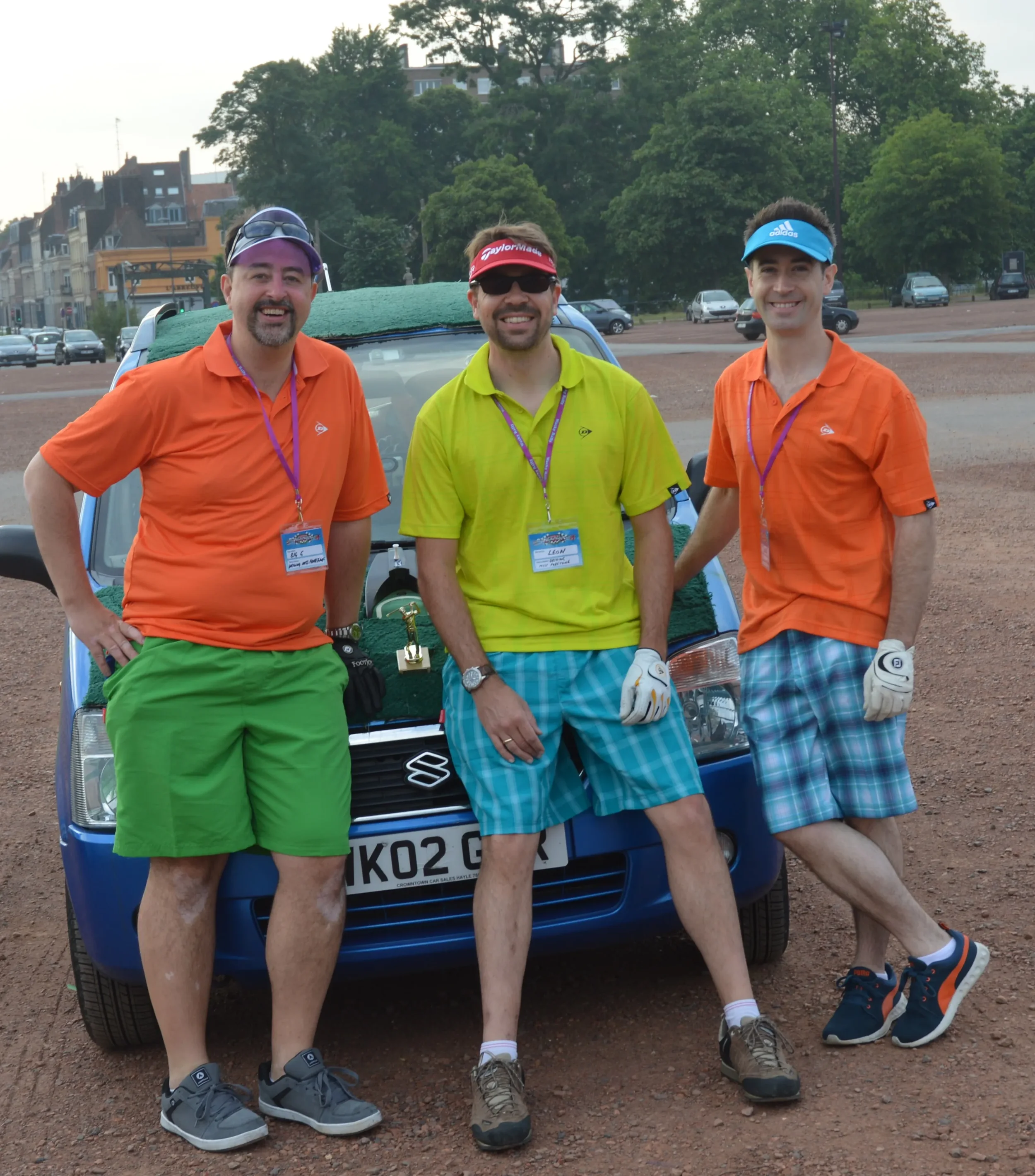 Three men standing in front of a blue car with the trunk open, outdoors on a gravel surface, wearing colorful cycling or sports attire, with trees and buildings in the background.