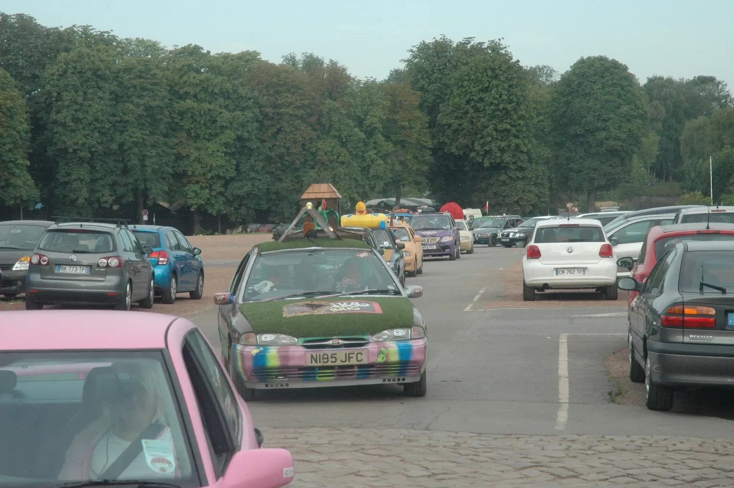 A parking lot with numerous cars, including a colorful, grass-covered car with a slide on its roof. Trees border the parking lot in the background.