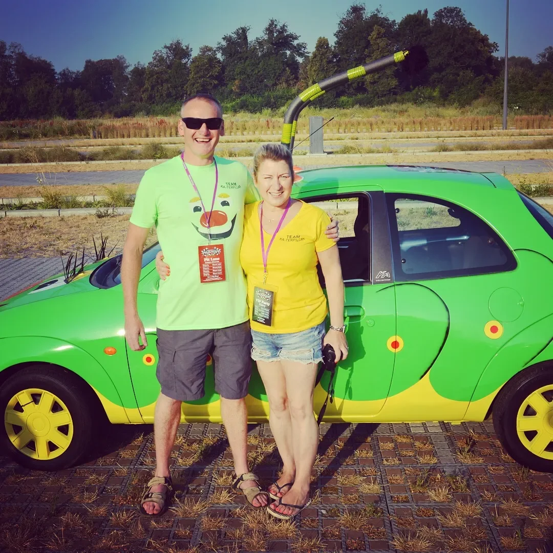 A man and a woman standing together in front of a bright green and yellow small car in an outdoor parking lot on a sunny day. The man is wearing sunglasses, a light green t-shirt with a smiling face graphic, gray shorts, and sandals. The woman is wea