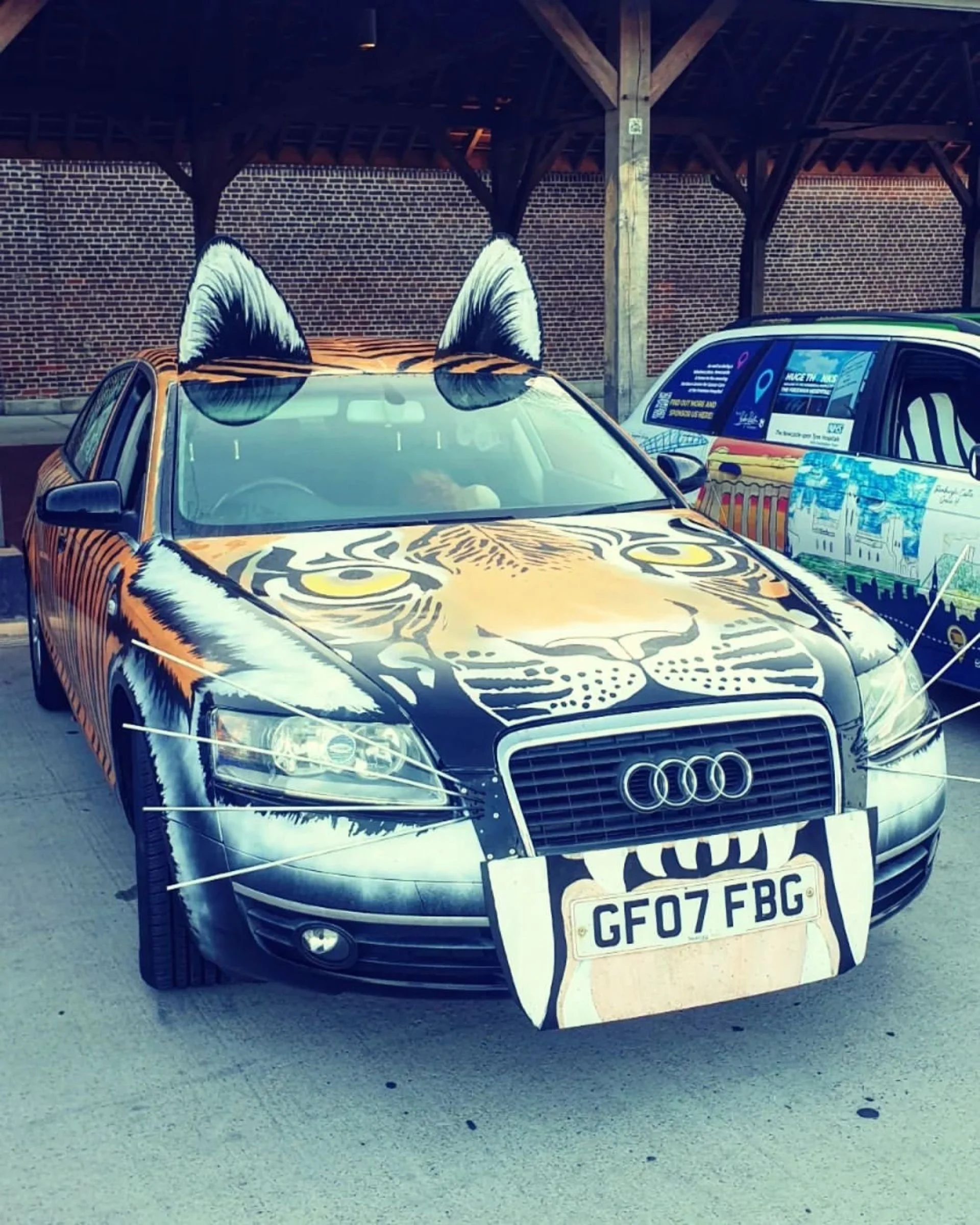 A black Audi car with tiger face and ears artwork on the hood and top, with a large tiger face design on the hood and tiger ears on the roof.