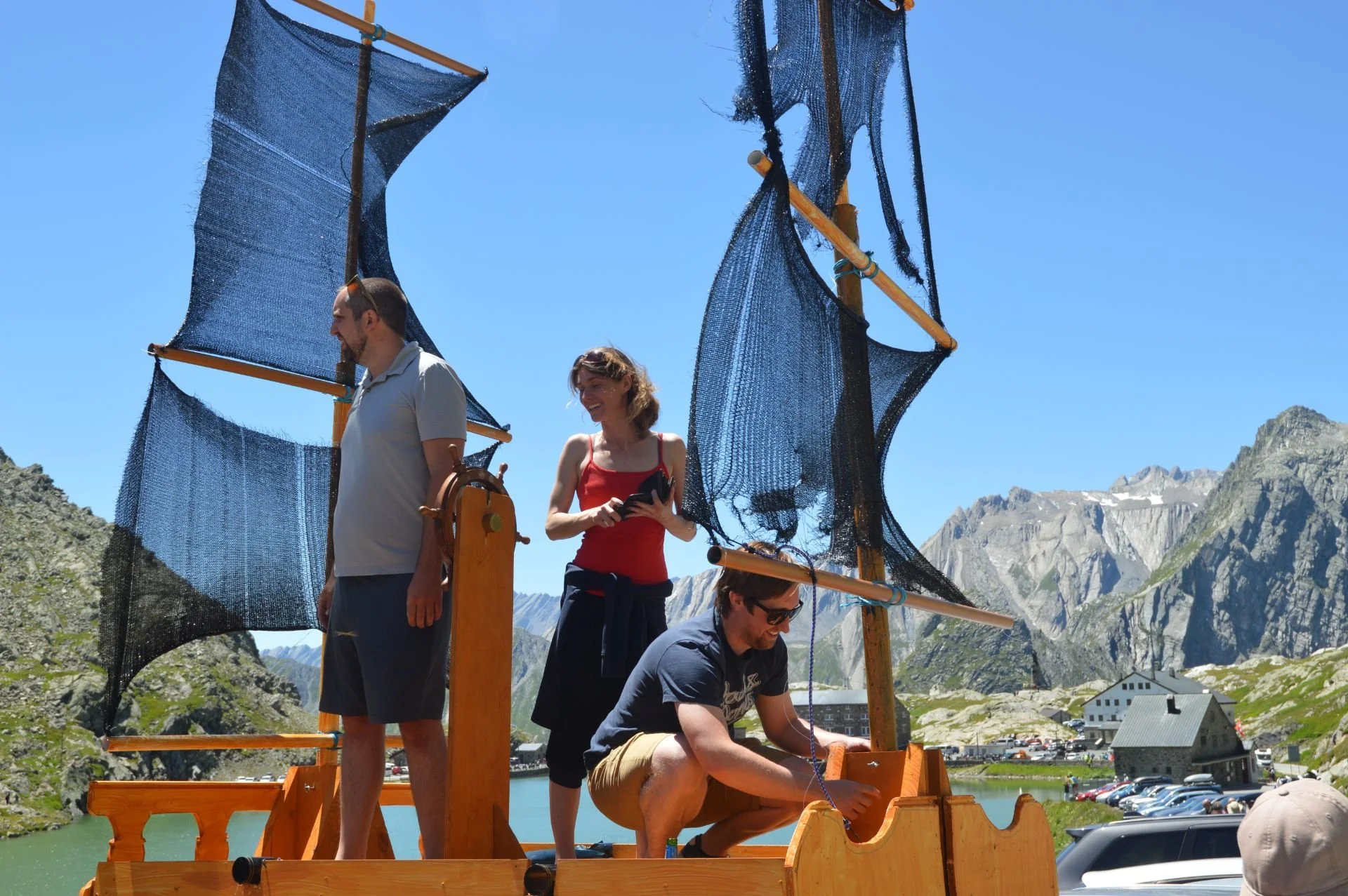 Three people on a small wooden boat resembling a ship, with large black sails made of mesh, set against a mountainous landscape with a small building and parking lot in the background, under a clear blue sky.