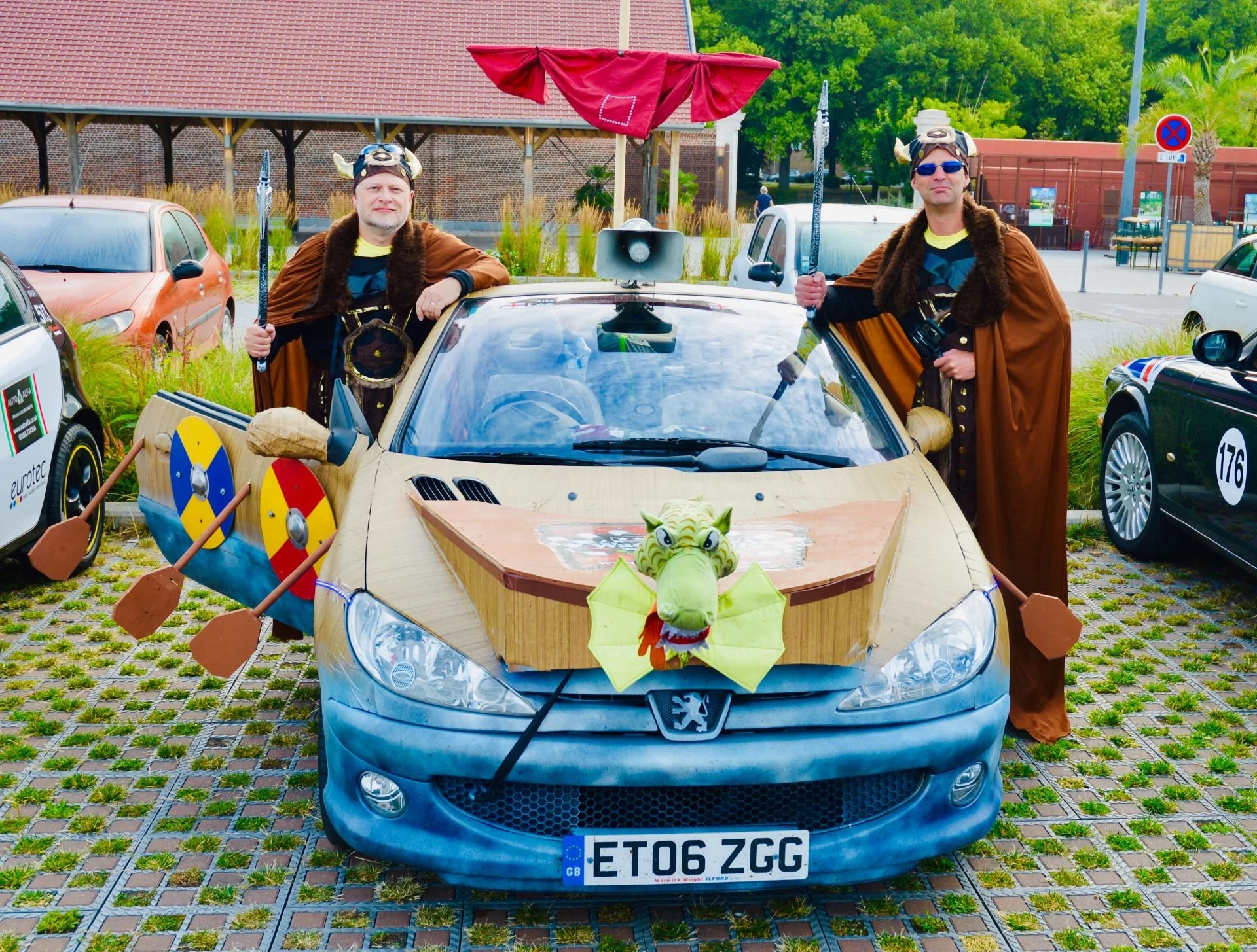Two men dressed as Vikings standing beside a decorated car with a dragon head figure, shields, and various props.