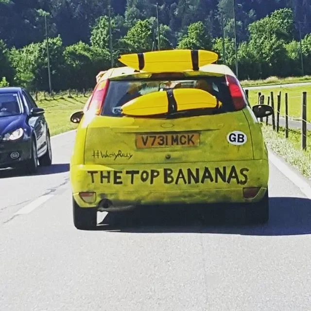 Yellow car with three yellow surfboards on top, a license plate V731 MCK, window sticker #WandJudy, and a humorous message painted on the back that says "THE TOP BANANAS".