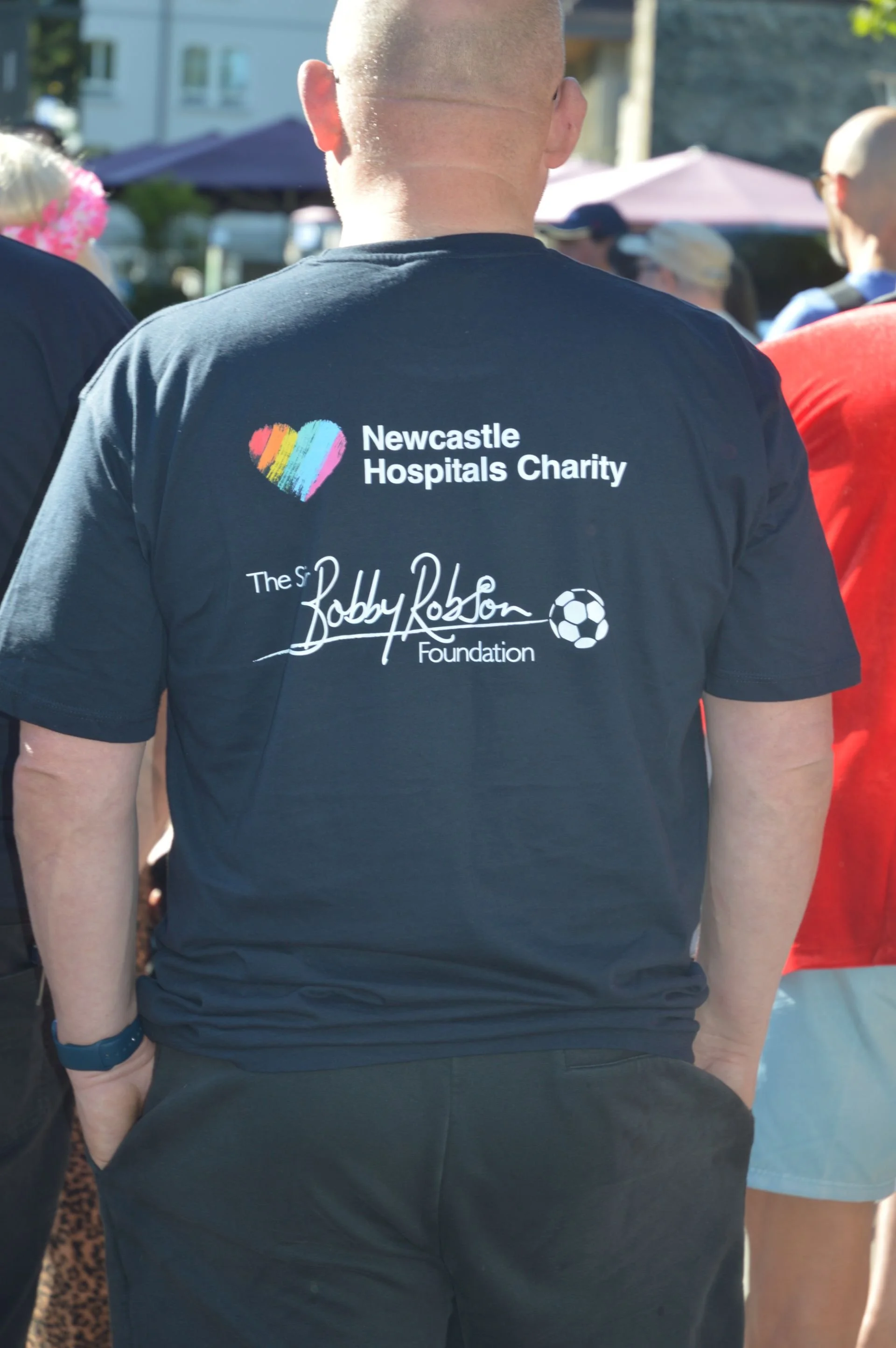 Back of a person wearing a black T-shirt with logos for Newcastle Hospitals Charity and the Bobby Robson Foundation, standing outdoors during daytime.