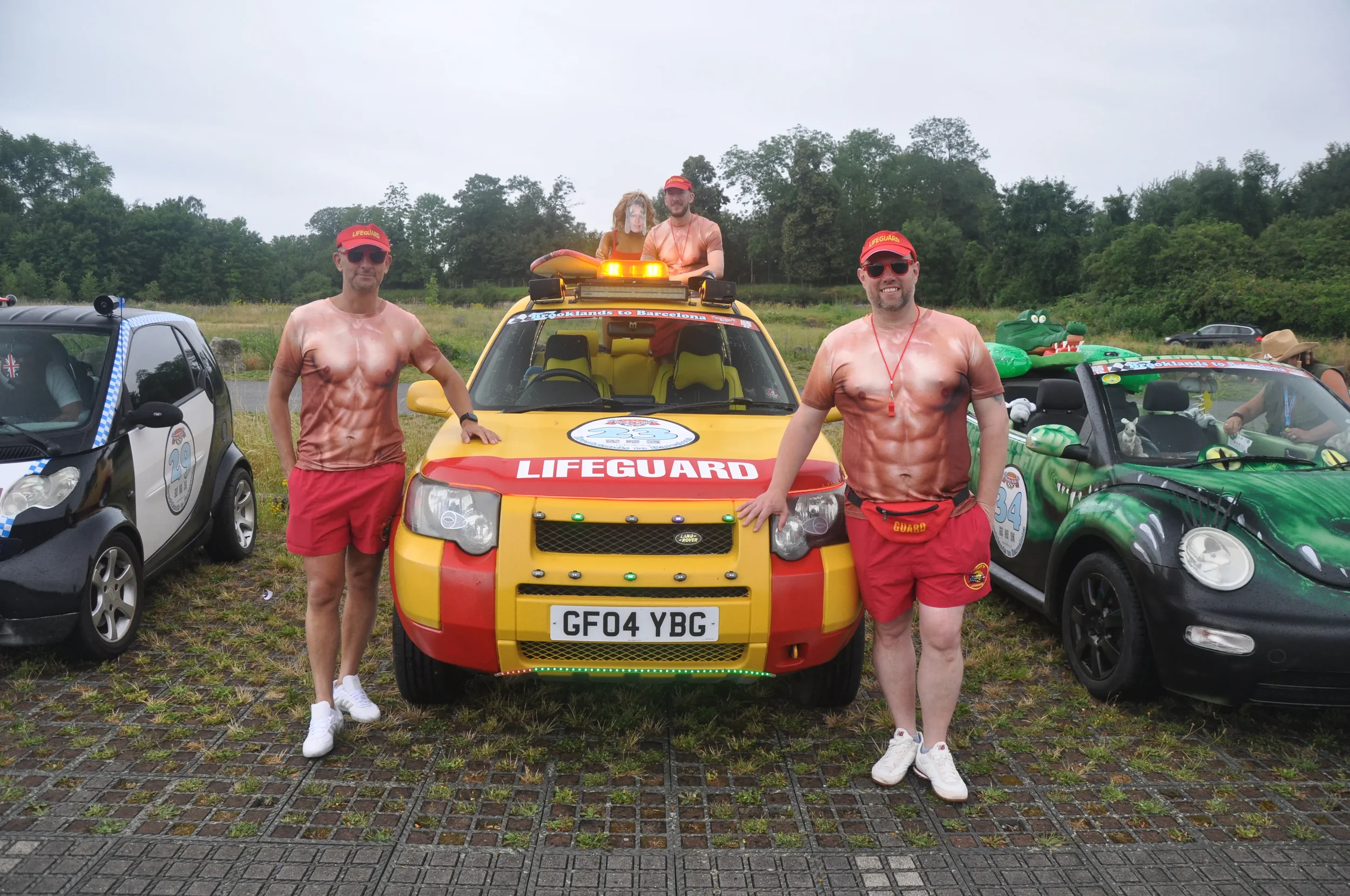 Three men dressed as lifeguards in shorts and shirts with muscular print, standing in front of a yellow and red lifeguard vehicle, with two other decorated cars on either side, outdoors on a grassy area.