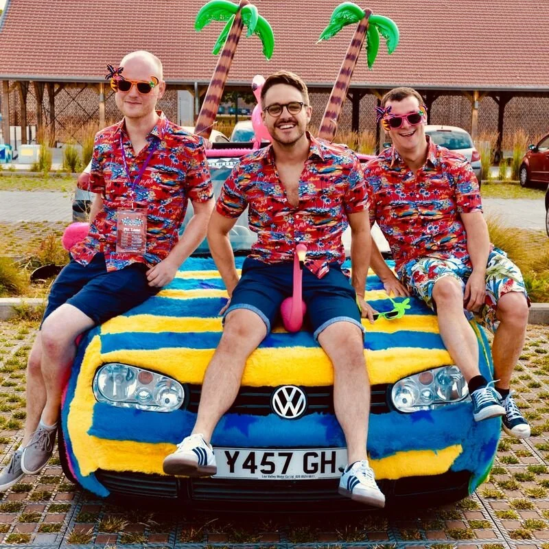 Three men in Hawaiian shirts sitting on the hood of a colorful car decorated with inflatable palm trees, flamingo, and swim floats at an outdoor event.