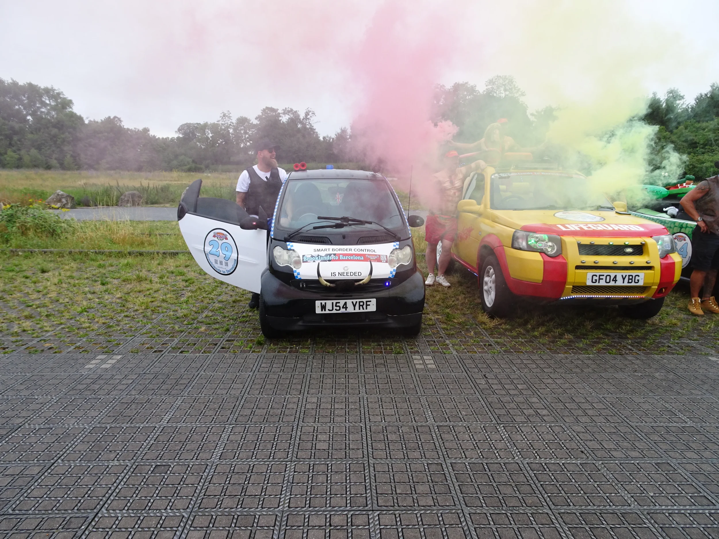 Two small vehicles, one black and one yellow with red accents, are parked outdoors during daytime. The black vehicle has an open door and a person standing nearby, while the yellow vehicle emits green and pink smoke. Several people are around the veh