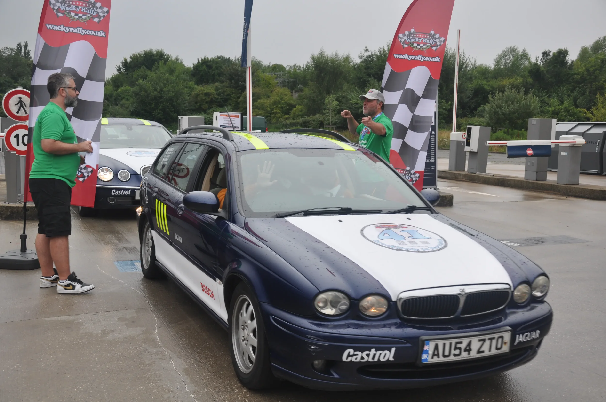 A blue and white vintage Jaguar racing car with the number 41 on the hood is parked at a rally event, with two men in green shirts standing nearby, one of whom is holding papers and the other is gesturing. There are checkered flags and rally banners 