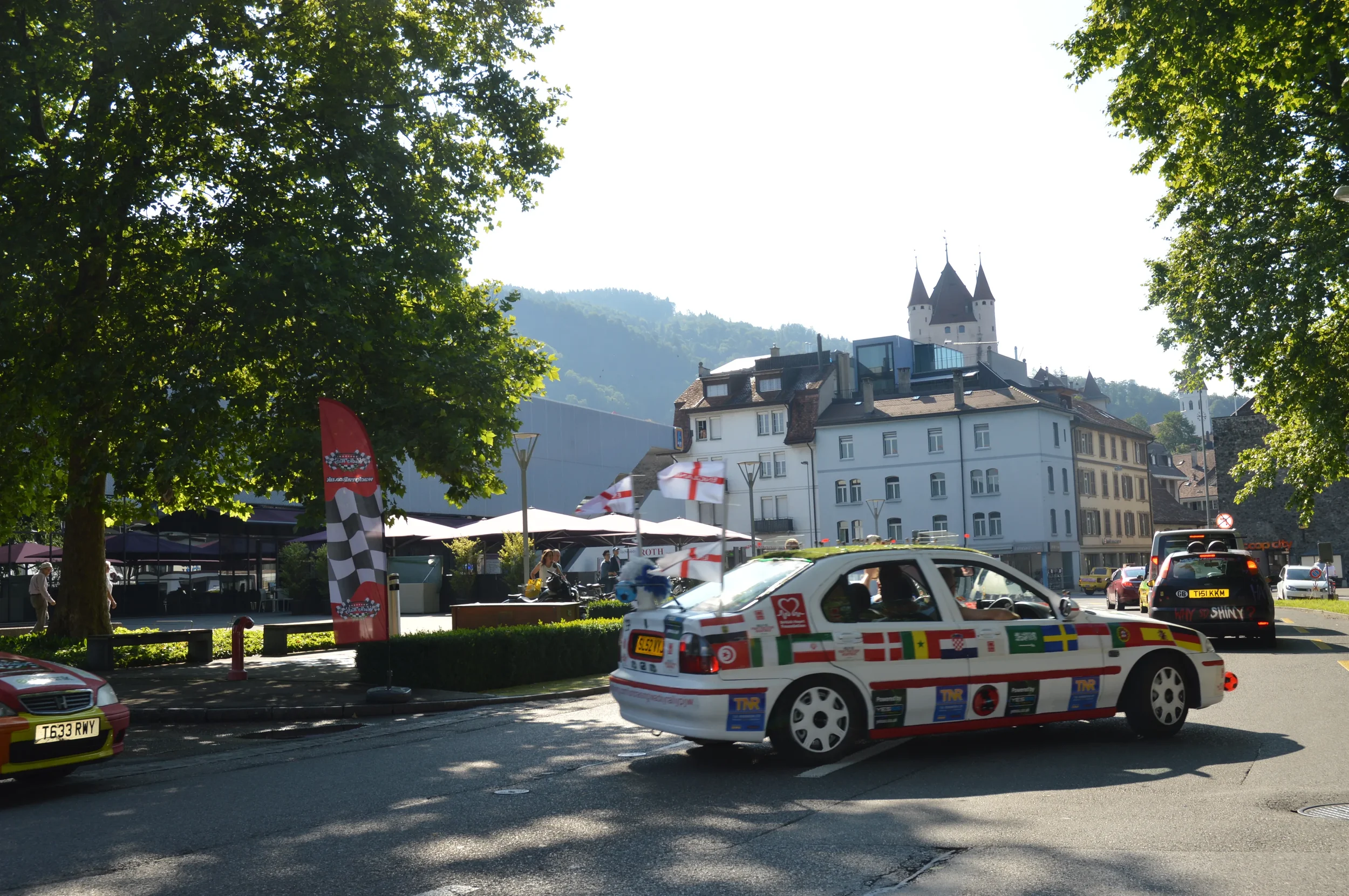 A white race car decorated with various national flags and sponsor stickers driving on a city street, with trees and historic buildings, including a castle, in the background.