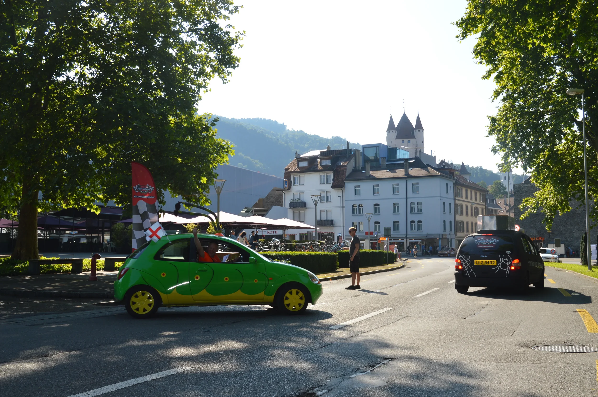 A colorful small green car with yellow wheels parked on a street in a town, with a person waving from the window, standing next to a person in black standing on the street. There are trees, buildings, and a castle in the background, with a mountain b