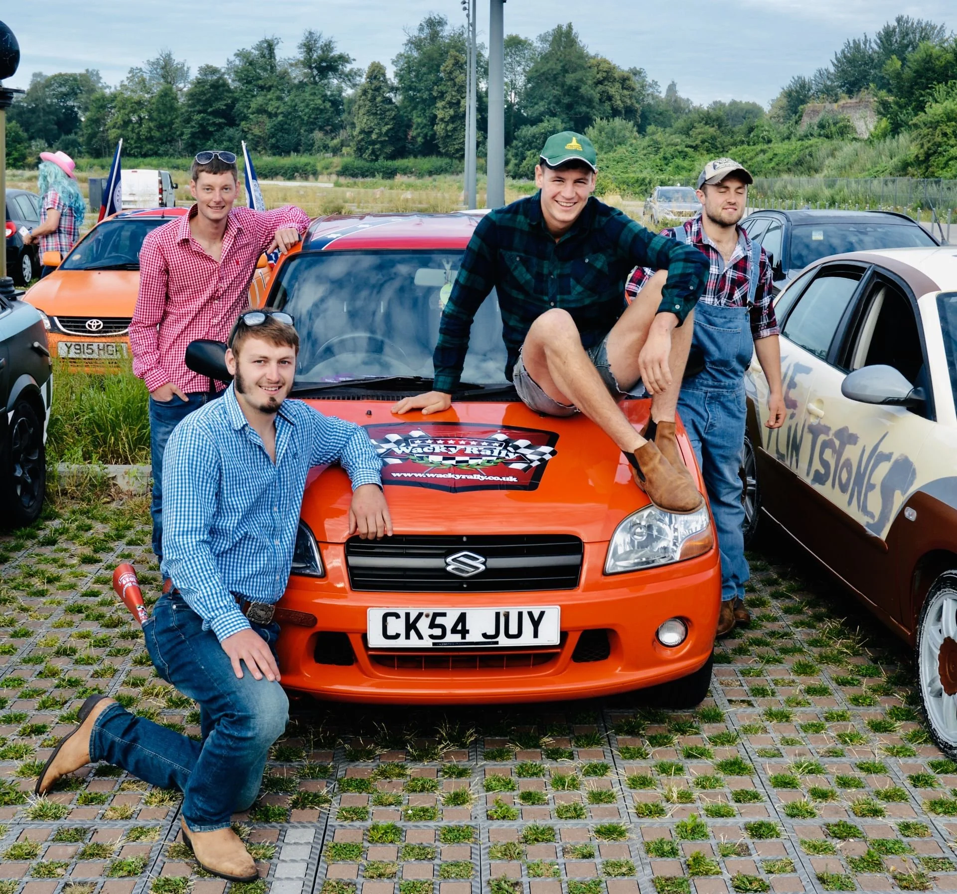 Group of five young men posing around an orange Suzuki car at an outdoor event, with some wearing plaid shirts and jeans, one sitting on the car's hood, and others standing nearby. There are other cars and trees in the background.