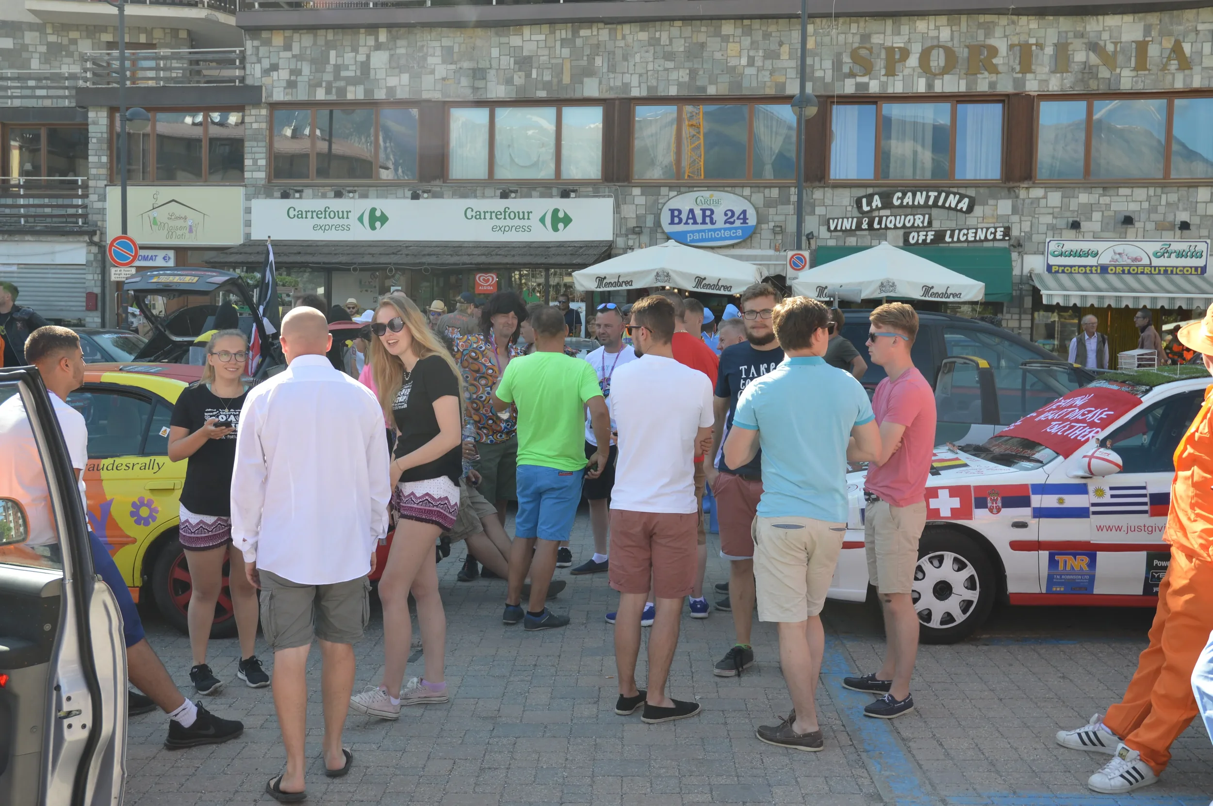 Group of people gathered in a street with decorated cars, including a car with Swiss and Greek flags, in front of shops and cafes.