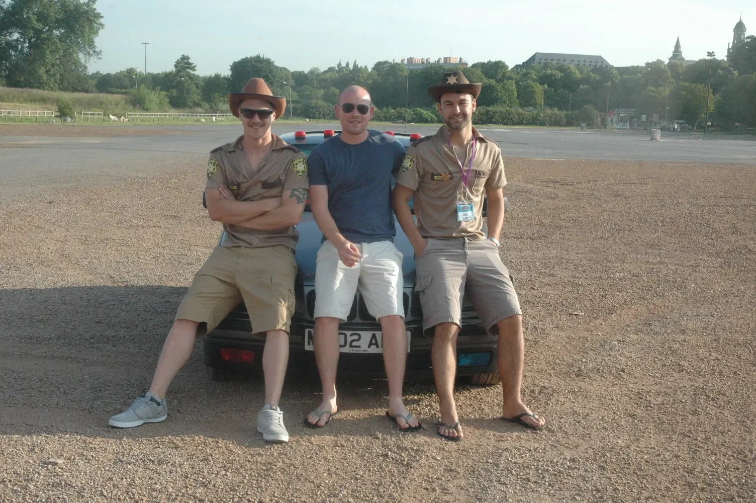 Three men sitting on the front of a police car in an open area, two dressed as police officers and one in casual clothing, with greenery and distant buildings in the background.