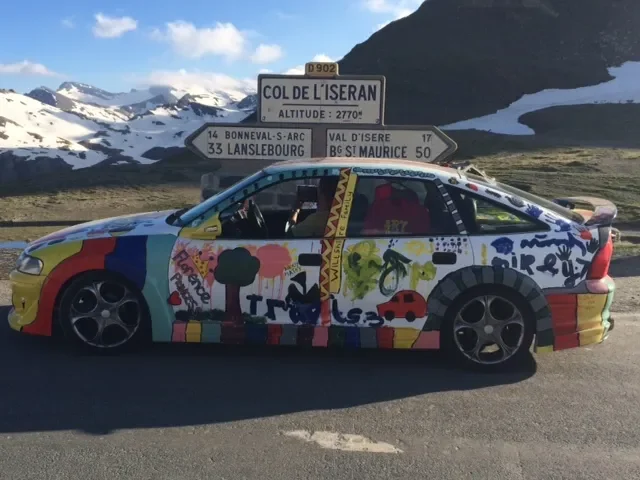 A colorful car decorated with various paint designs parked next to road signs indicating locations and altitude, with mountains and snow in the background.