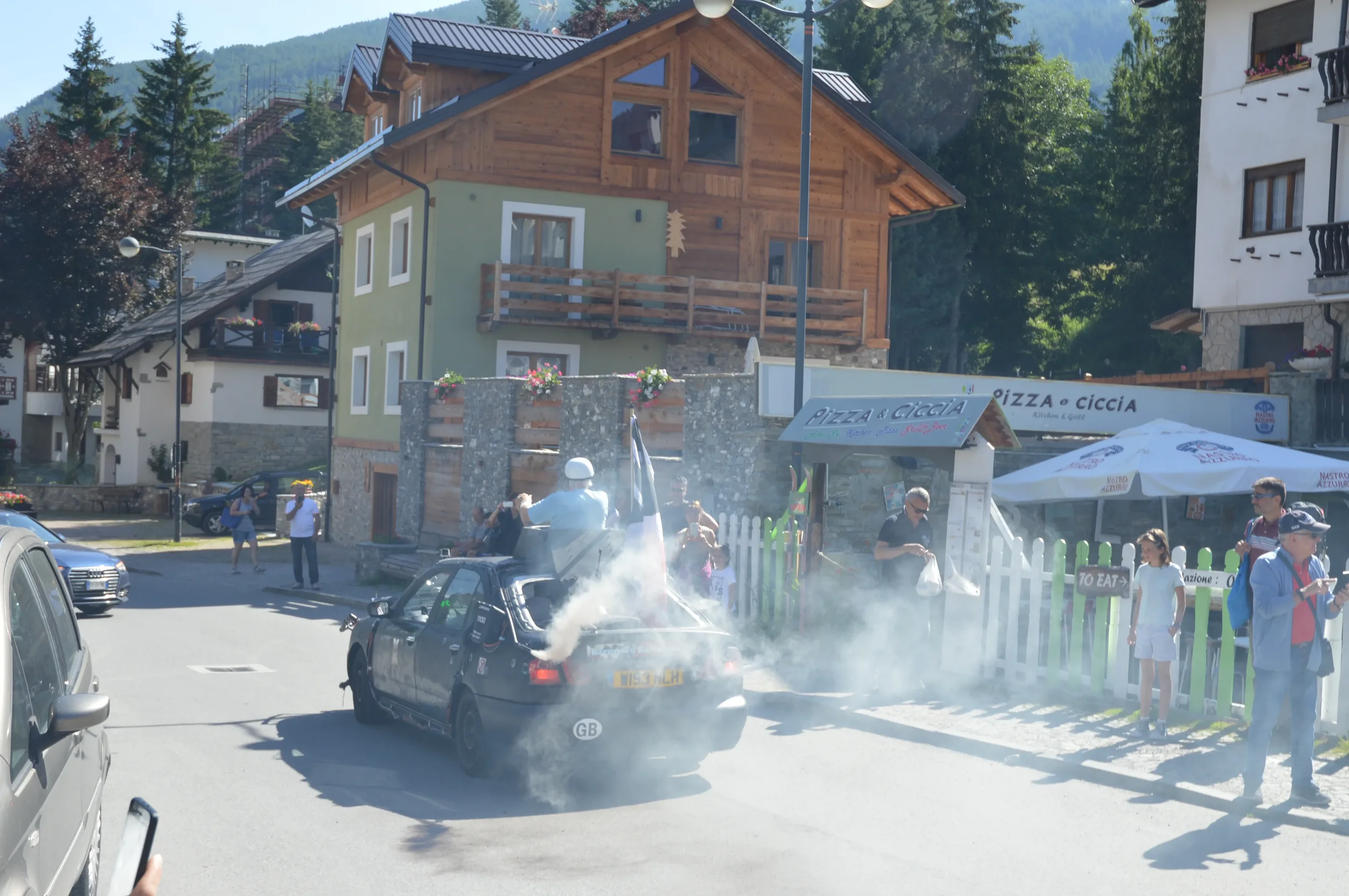 A street scene in a mountain town with a small crowd gathered around a black car emitting smoke or steam. Some people are taking photos or looking at the scene, with buildings and trees in the background.