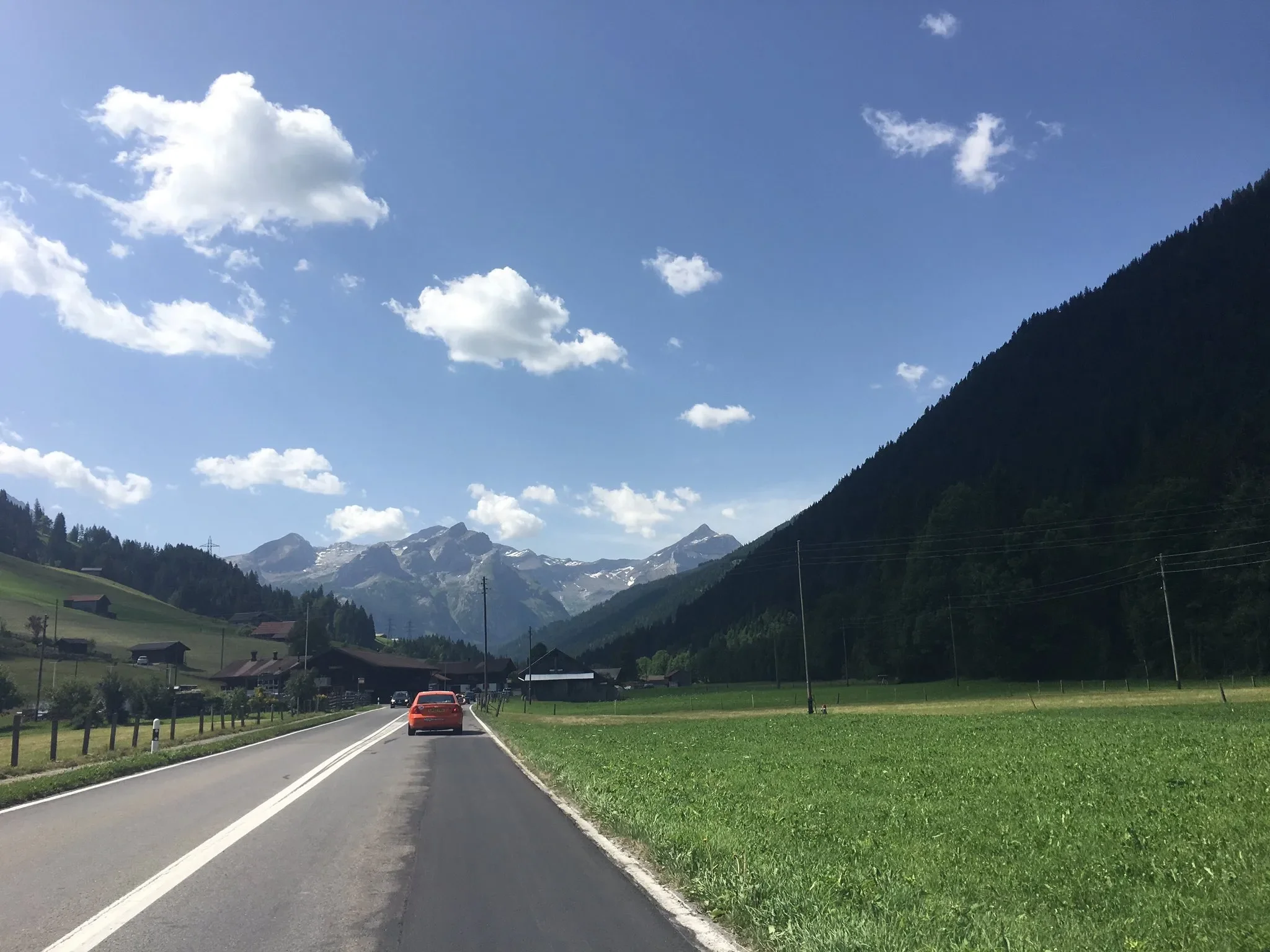 A scenic mountain landscape with a two-lane road, a red car, green fields, houses, and snow-capped mountains in the distance under a partly cloudy sky.