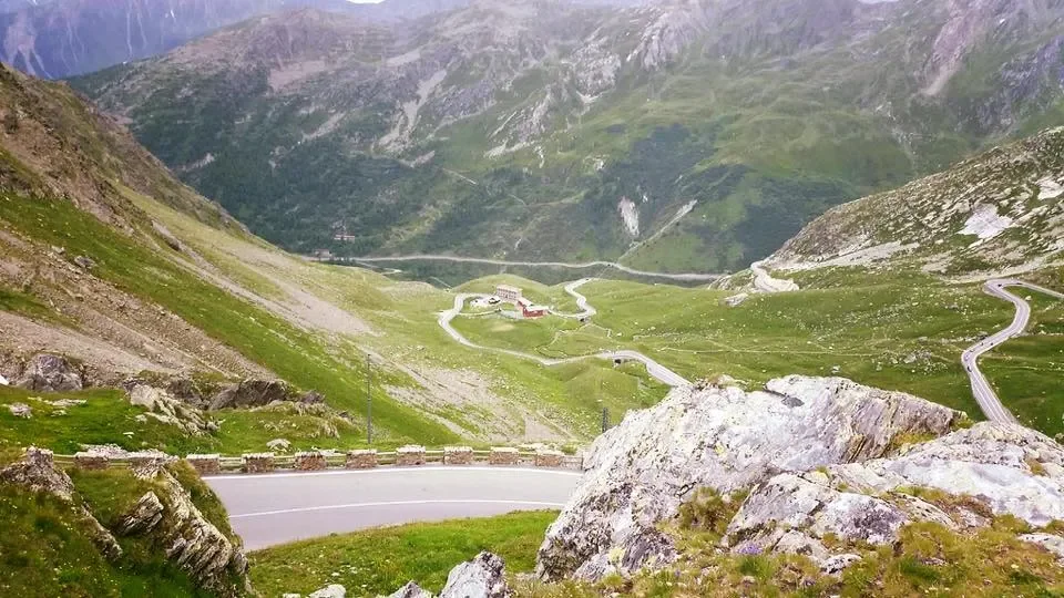 A winding road through a mountainous landscape with green grass and rocks, leading towards distant buildings in a valley.