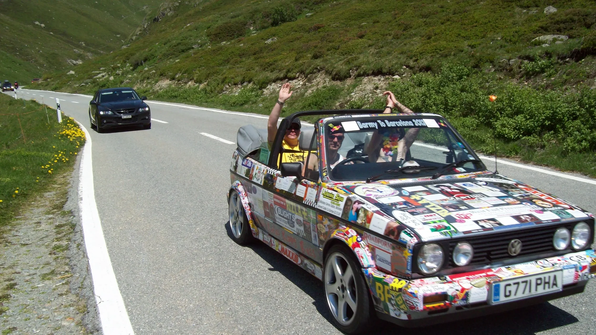 A colorful Volkswagen convertible with stickers and decals, carrying three people, driving on a mountain road with cars ahead, waving at the camera.