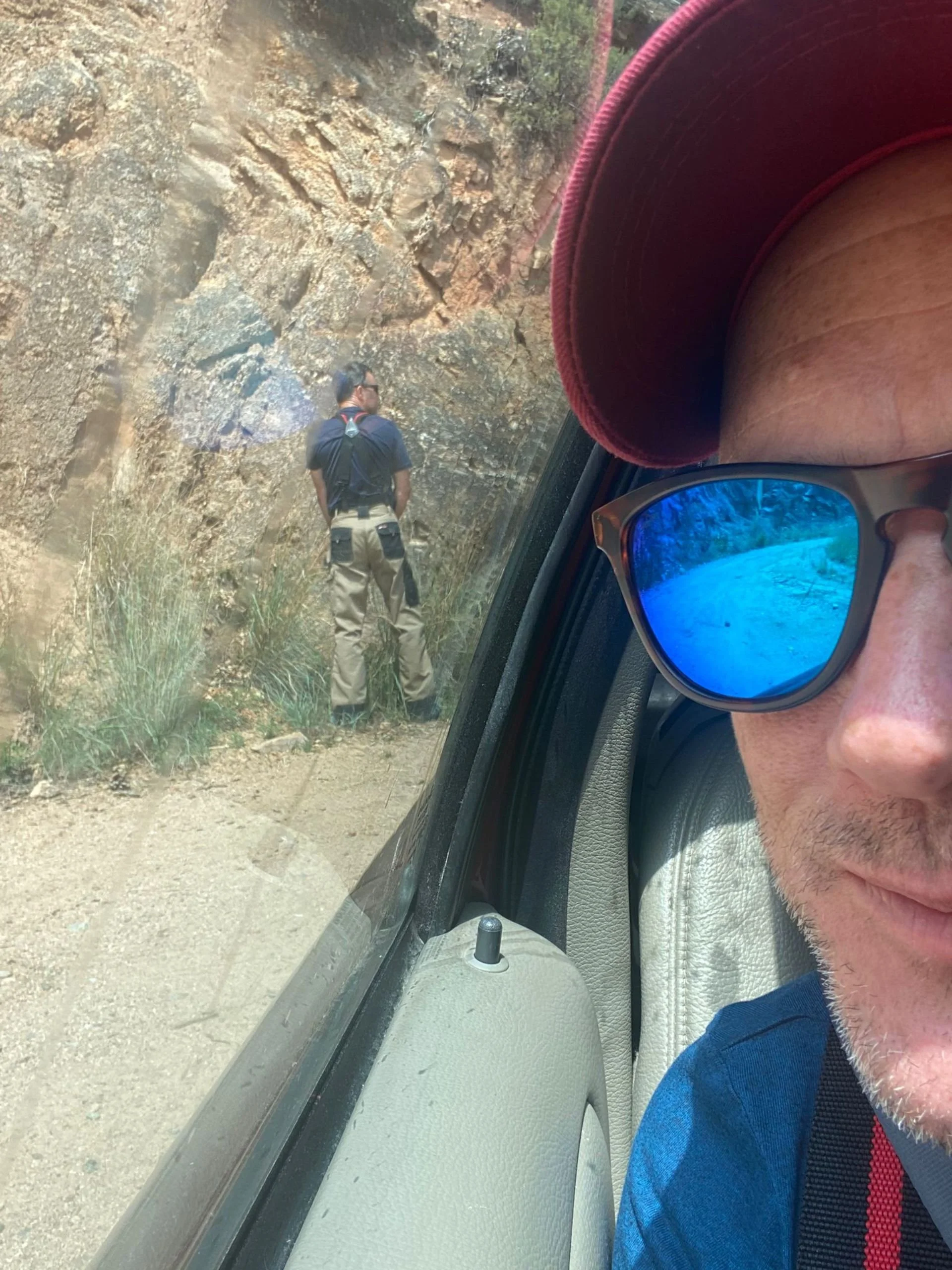 Person in sunglasses and a baseball cap inside a vehicle taking a selfie, with a man standing outside the vehicle on a dirt trail near a rocky hillside.